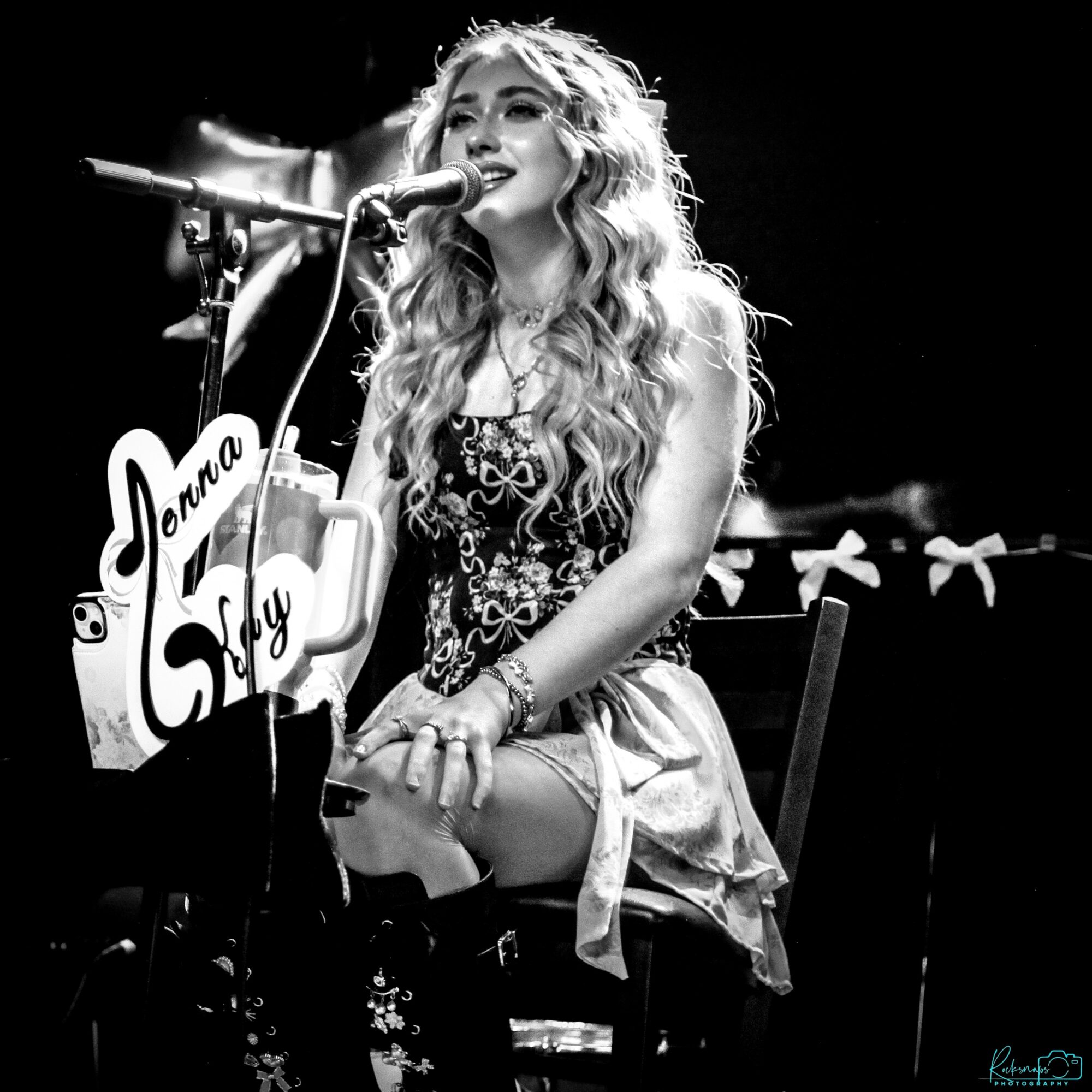 Young woman with long curly hair singing into a microphone on stage, sitting on a chair, with a sign reading 'Danna Sky'.