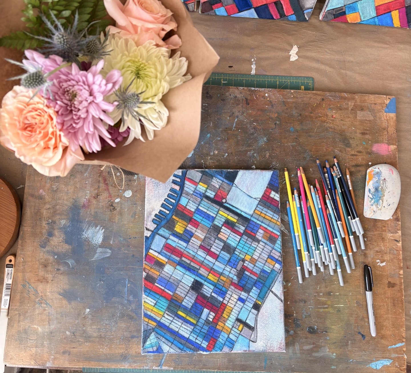 Colorful mosaic pattern on paper, surrounded by markers, a ruler, and a flower bouquet on a wooden table.