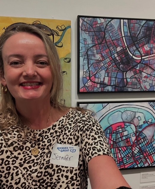 Smiling woman with blonde hair wearing a leopard print top and name tag, standing in front of city maps.