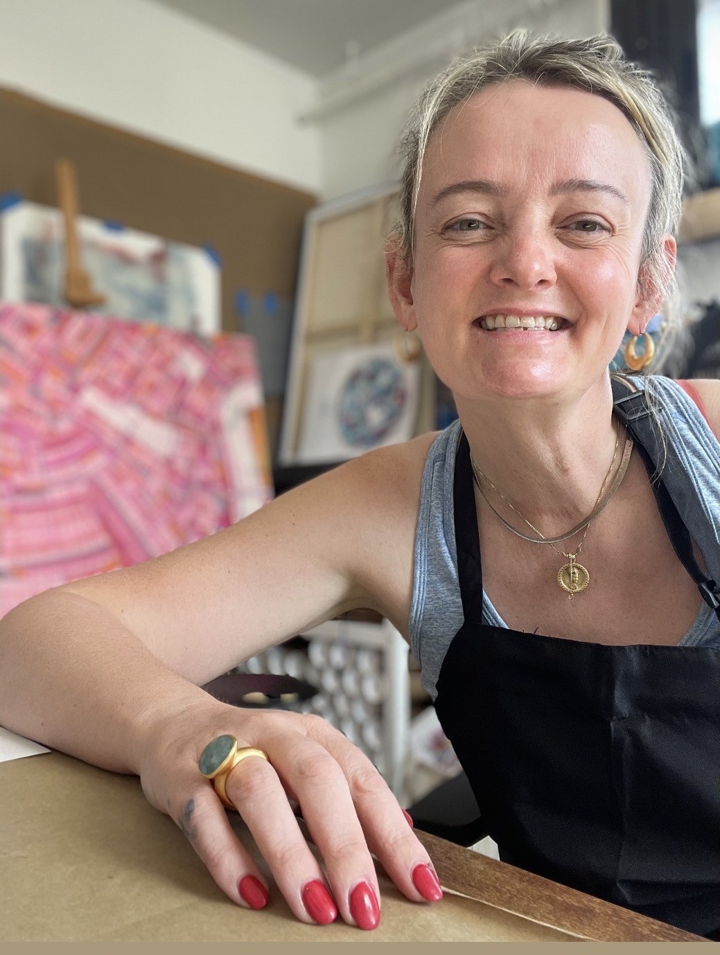 Smiling woman with gray hair, wearing a black apron and necklace, sitting at a table in an art studio.