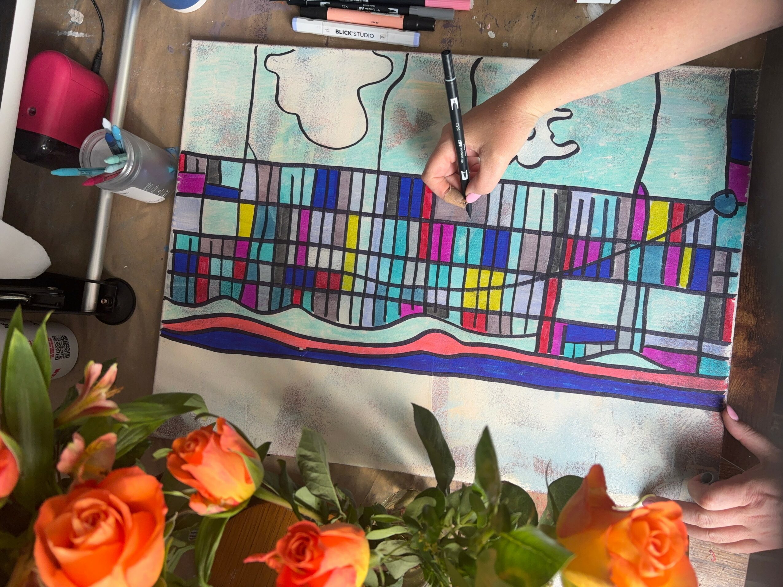Person coloring a large, colorful abstract drawing on paper with markers, surrounded by flowers and desk items.
