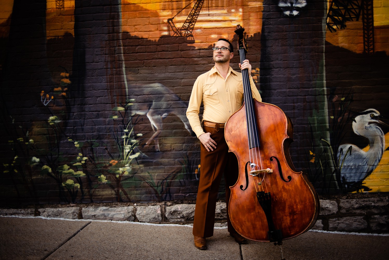 Man standing outdoors with a large double bass, mural background, sunset lighting, casual attire, hands on hips.