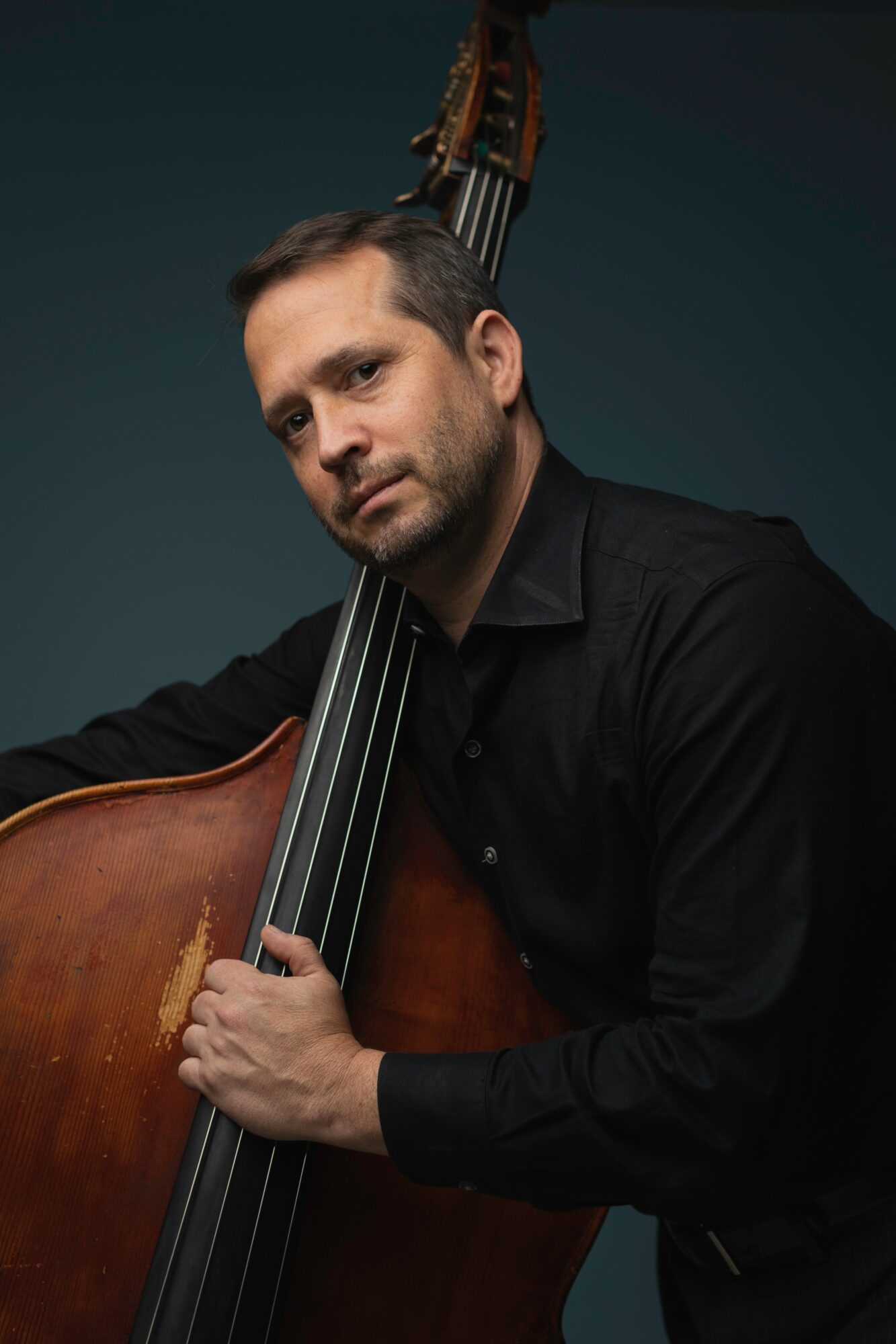 Man with short hair and beard holding a double bass, wearing a black shirt, against a dark background.