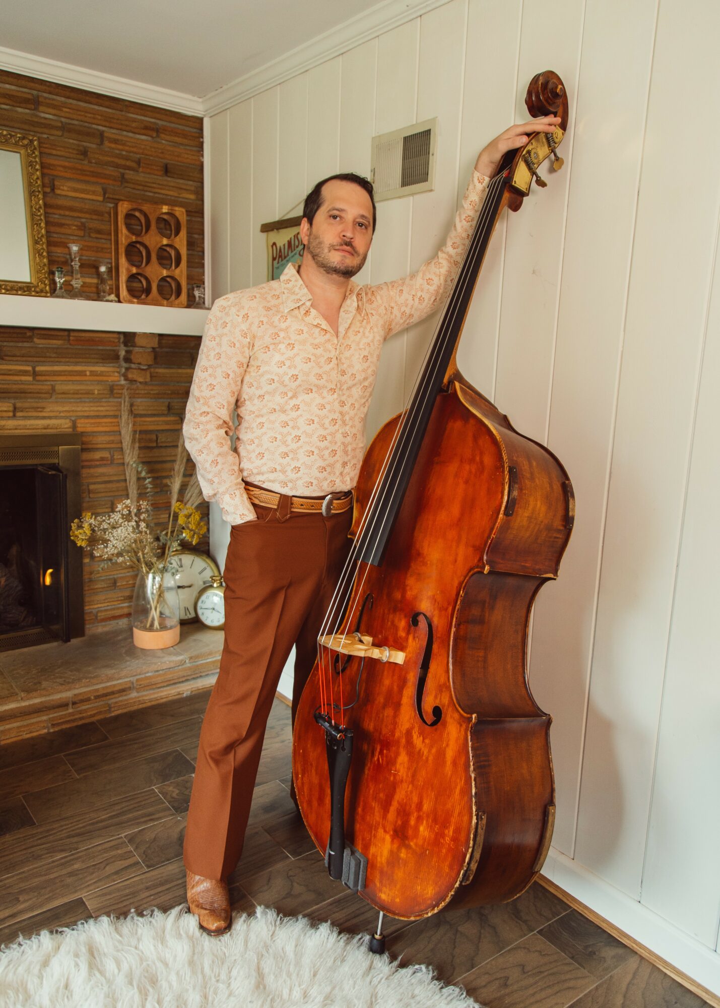 Man standing next to a large wooden double bass, touching its top, in a room with wood-paneled walls and fireplace.