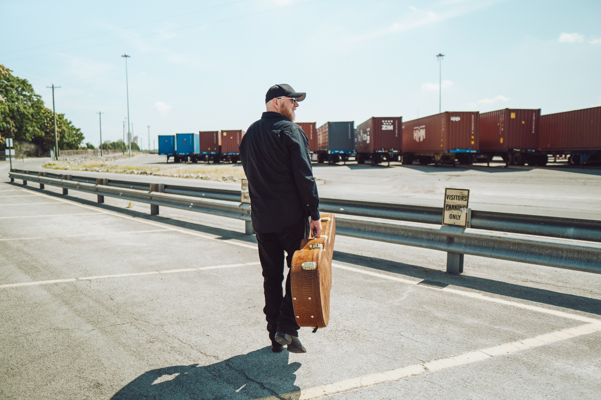 Person standing in a parking lot holding a suitcase, with train cars in the background, under a clear sky.