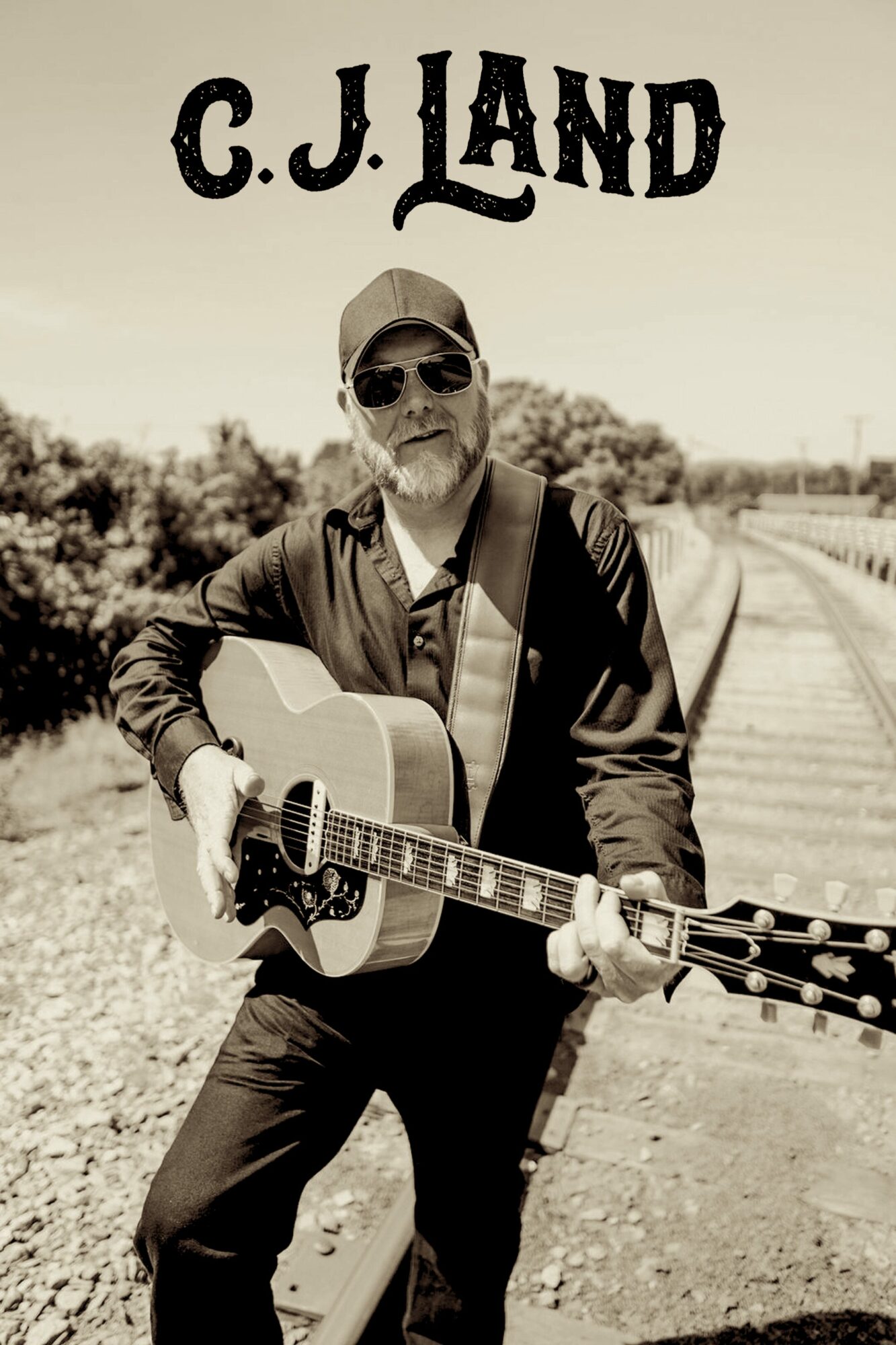 Man wearing sunglasses and a cap playing an acoustic guitar outdoors, with train tracks in the background.