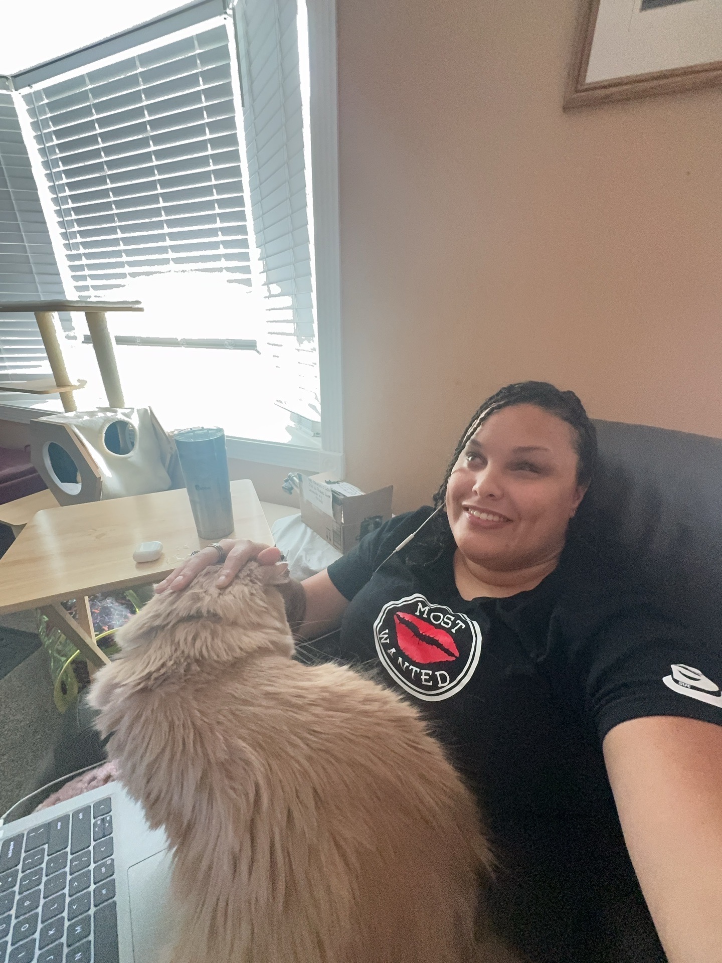 Person sitting in a chair smiling with a dog on their lap, in a room with a window and blinds.
