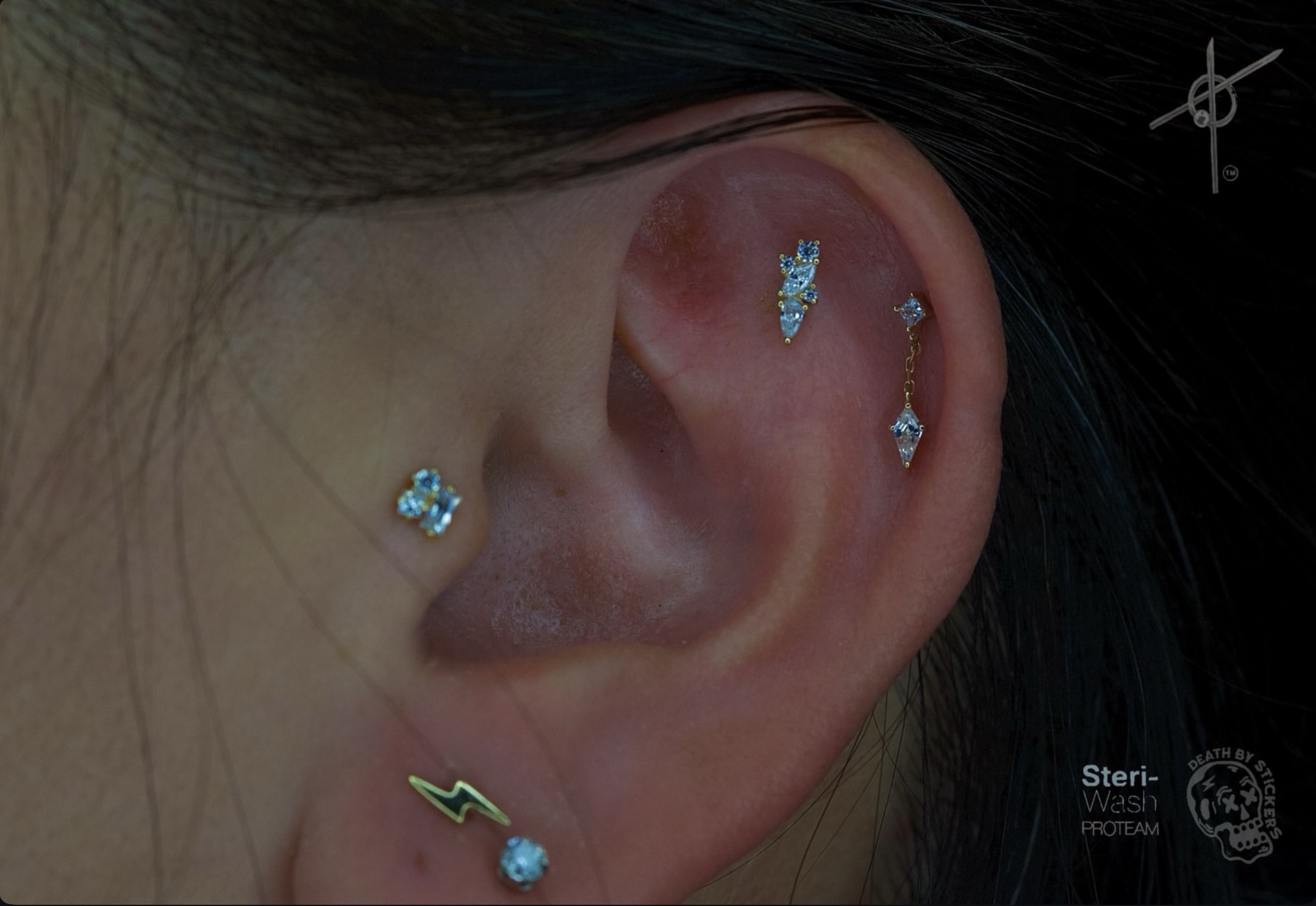 Close-up of a person's ear with multiple earrings, including a stud, a dangling earring, and a cuff, with dark hair.