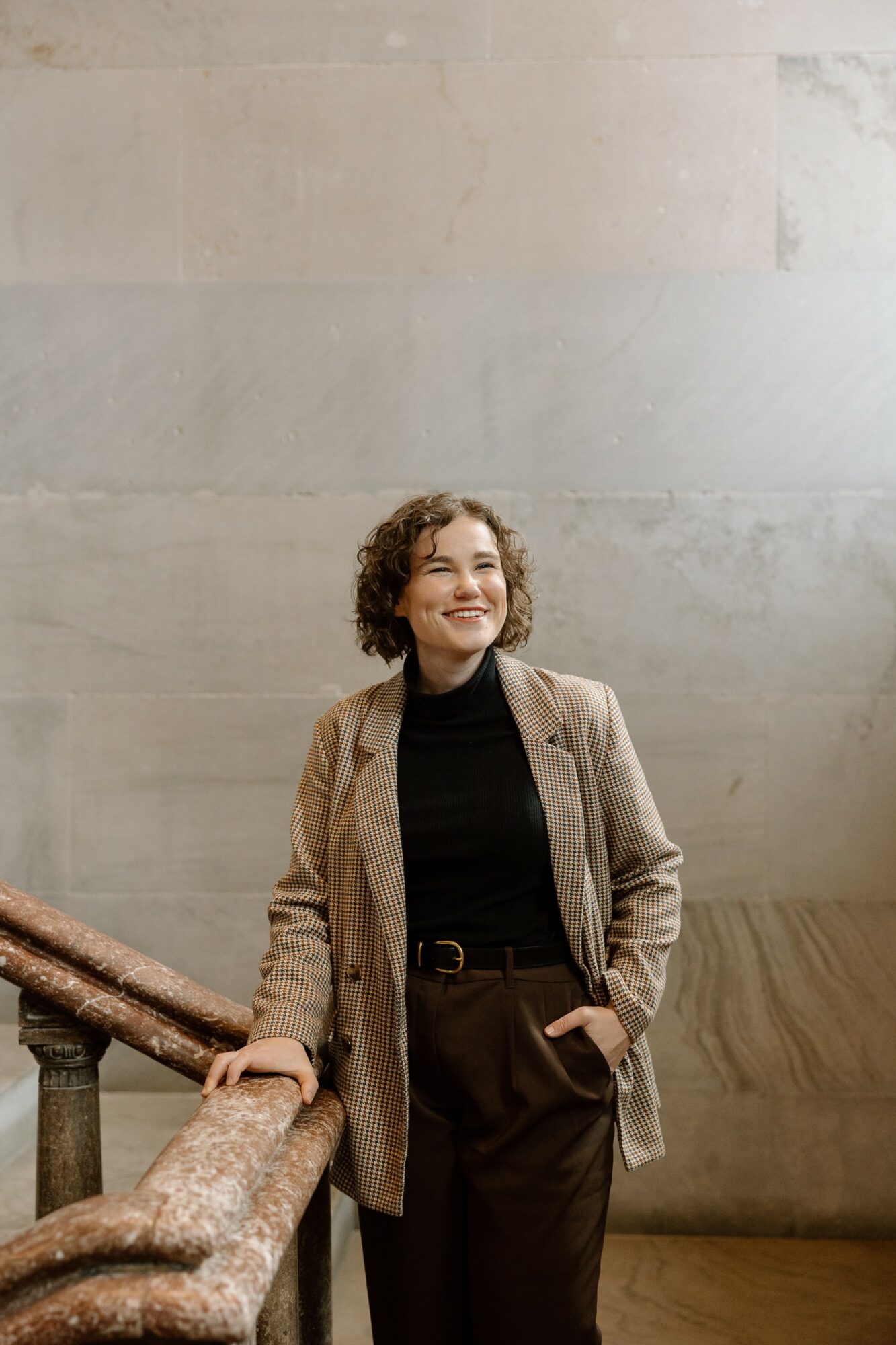 Woman with curly hair smiling, wearing a beige blazer and black outfit, standing near a railing against a plain wall.