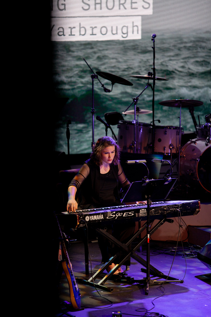 Musician playing keyboard on stage with drums and ocean background, with text partially visible in the top left corner.
