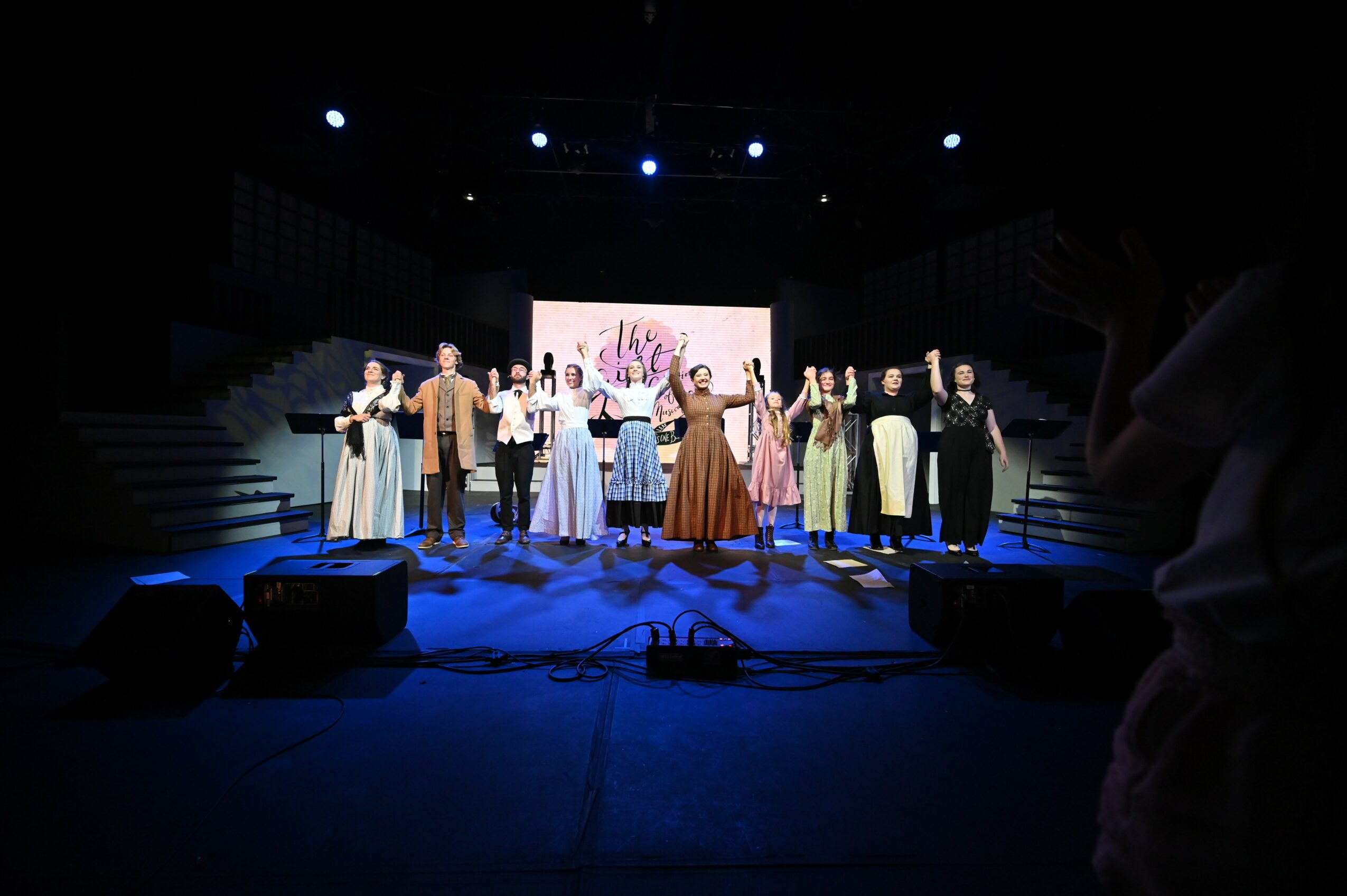 Group of people on stage holding hands, with a screen and stage lights in the background, in a dark theater setting.