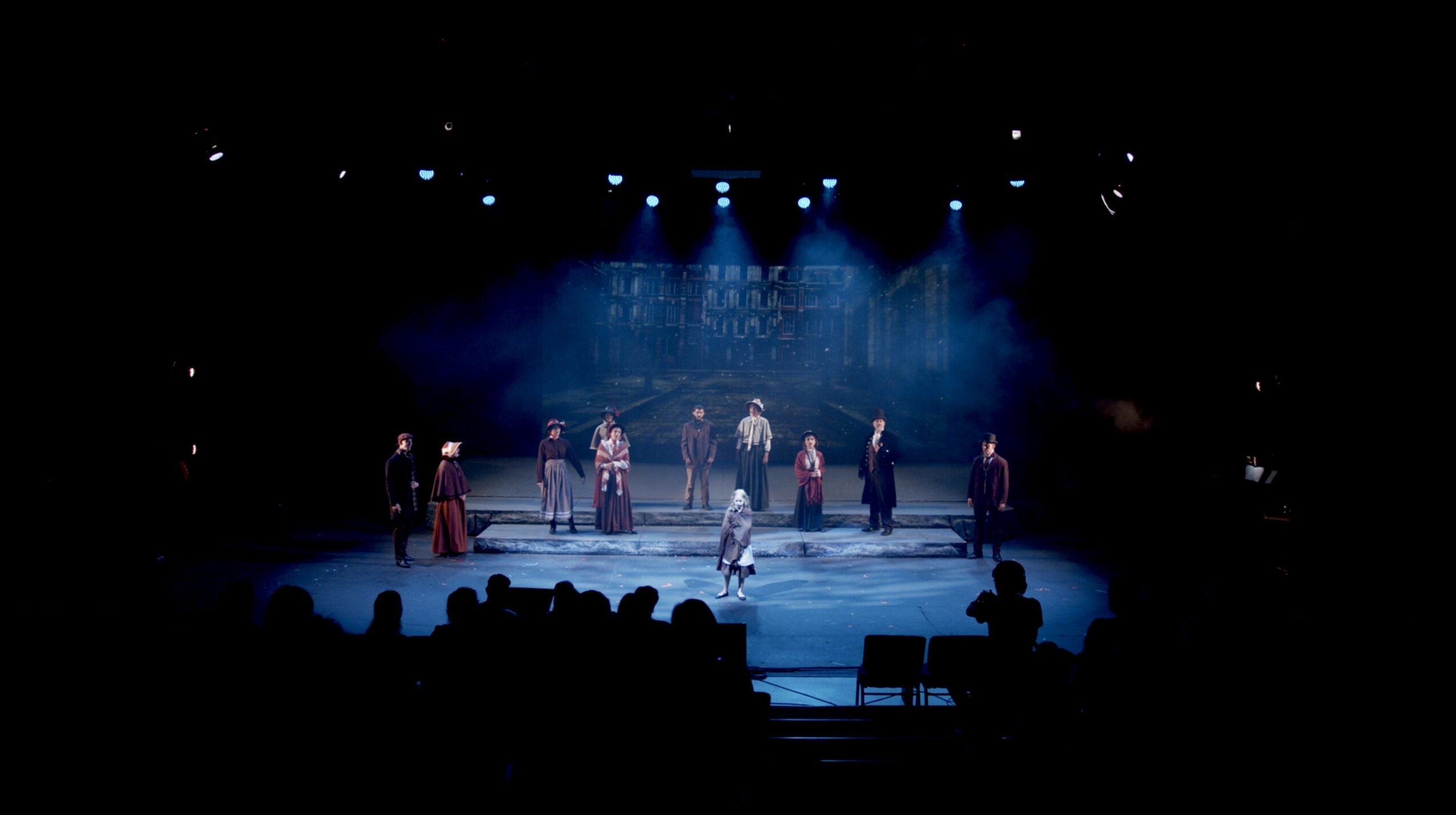Group of performers on stage with blue lighting, audience silhouettes visible, stage set in background.