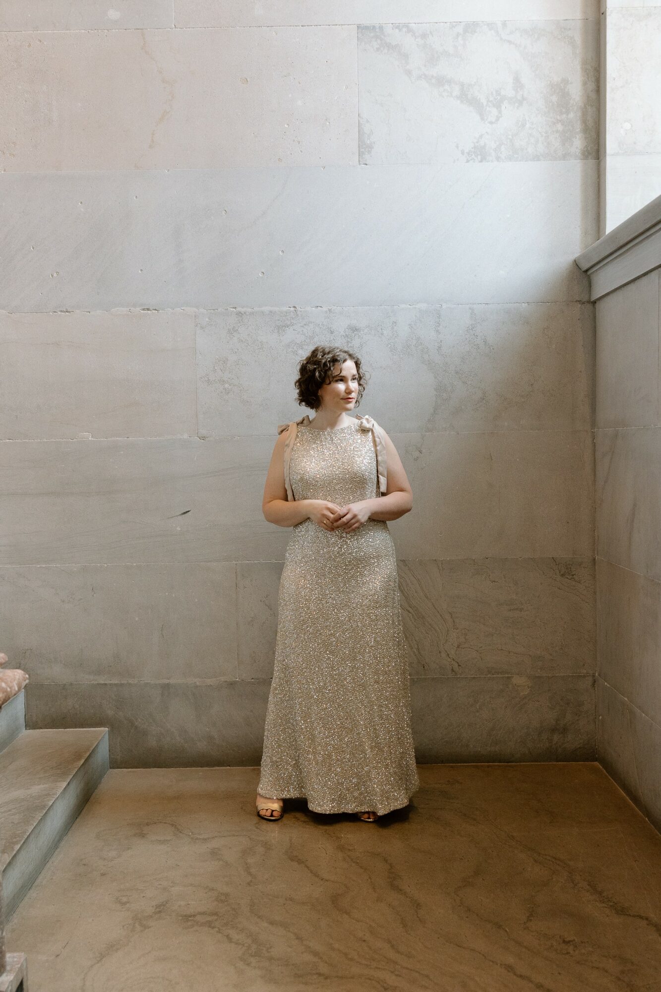 Woman in a long dress standing against a large, light-colored wall, looking to the side with hands clasped.
