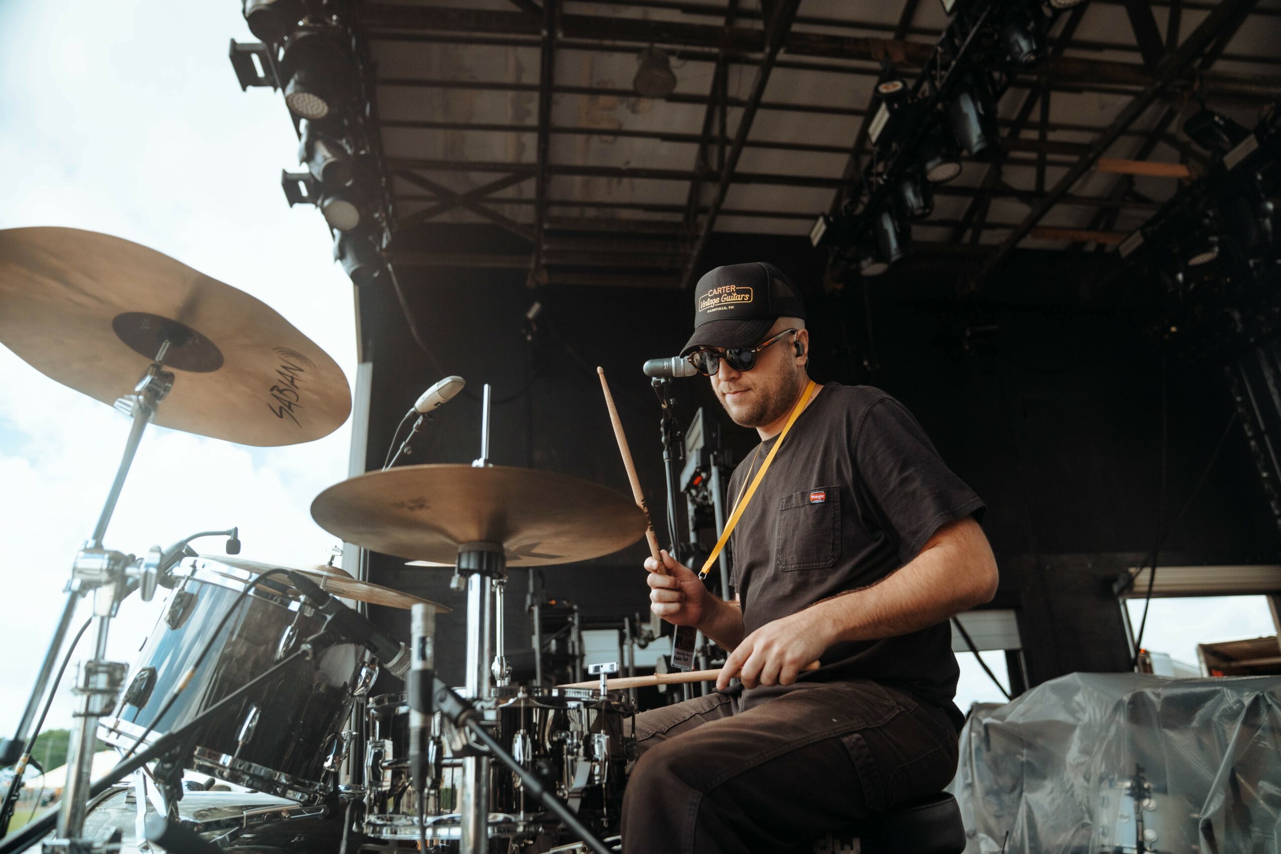 Person playing drums on stage with cymbals and drum set, wearing a cap and sunglasses, stage lighting above.