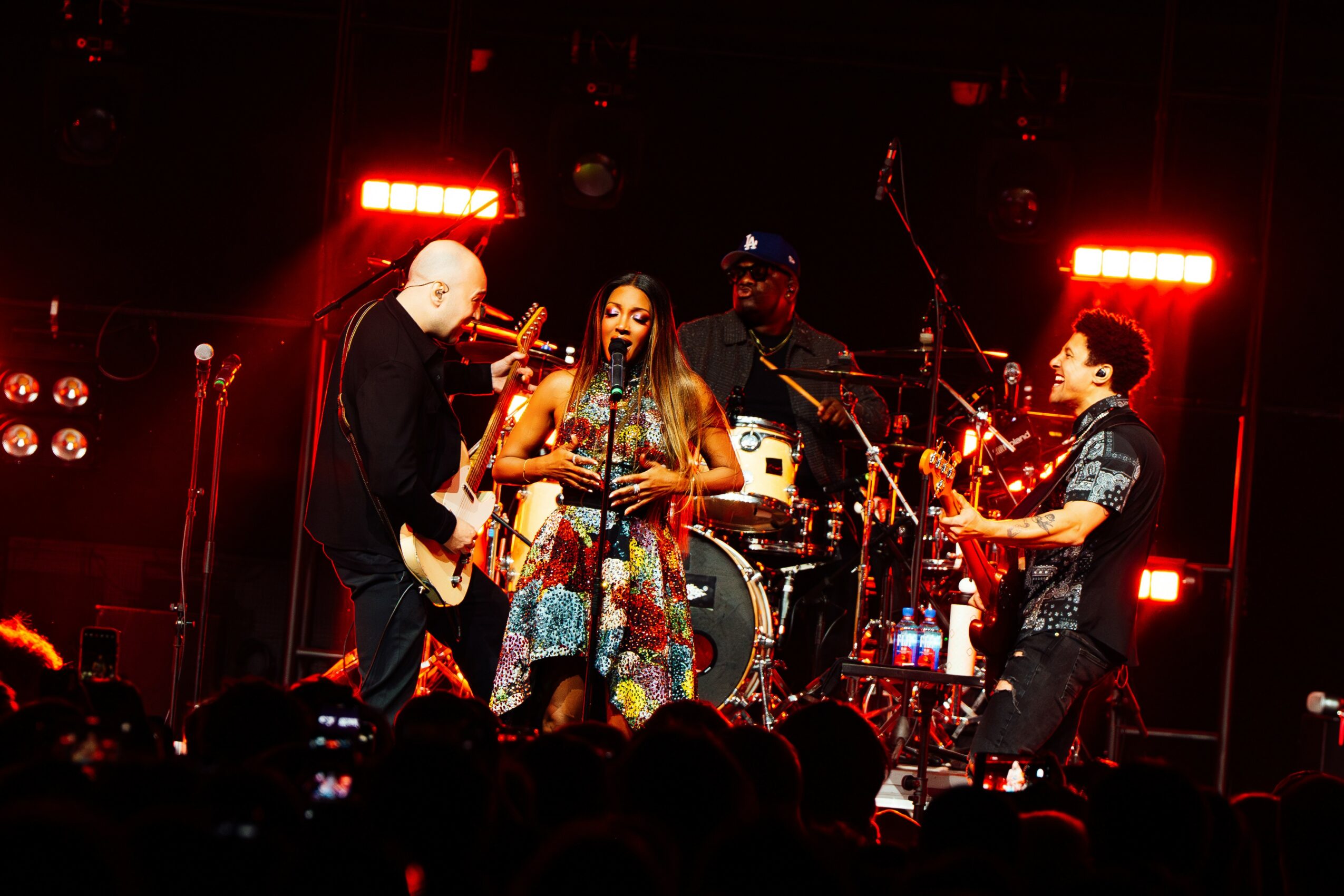 Four musicians perform on stage with red lighting, including a woman singing and playing guitar, and three men playing instruments.