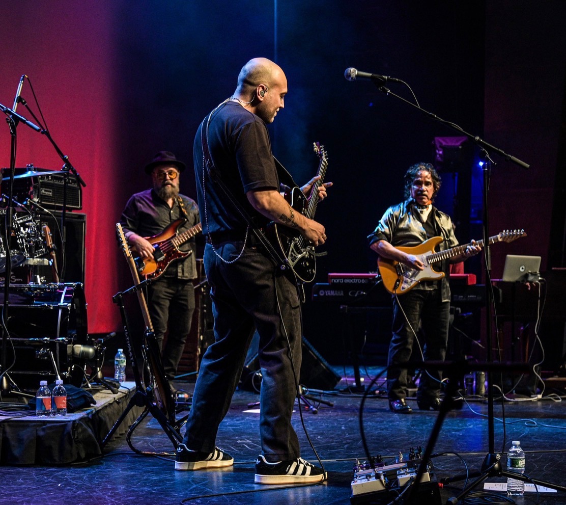Four musicians perform on stage with instruments, colorful lighting, and music equipment visible in the background.