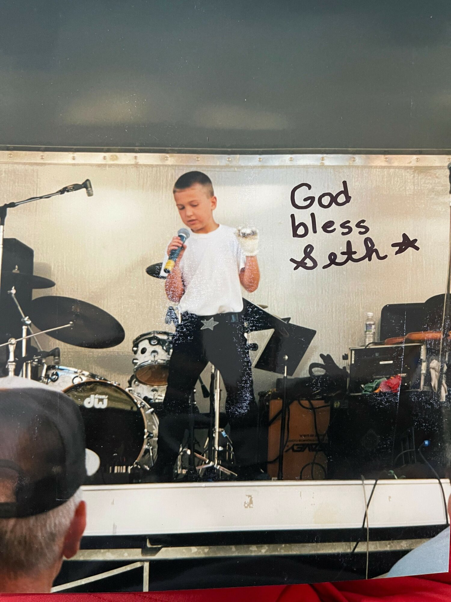 Person standing on stage with musical instruments, holding a microphone, with a sign that says 'God bless xBLACK' behind them.