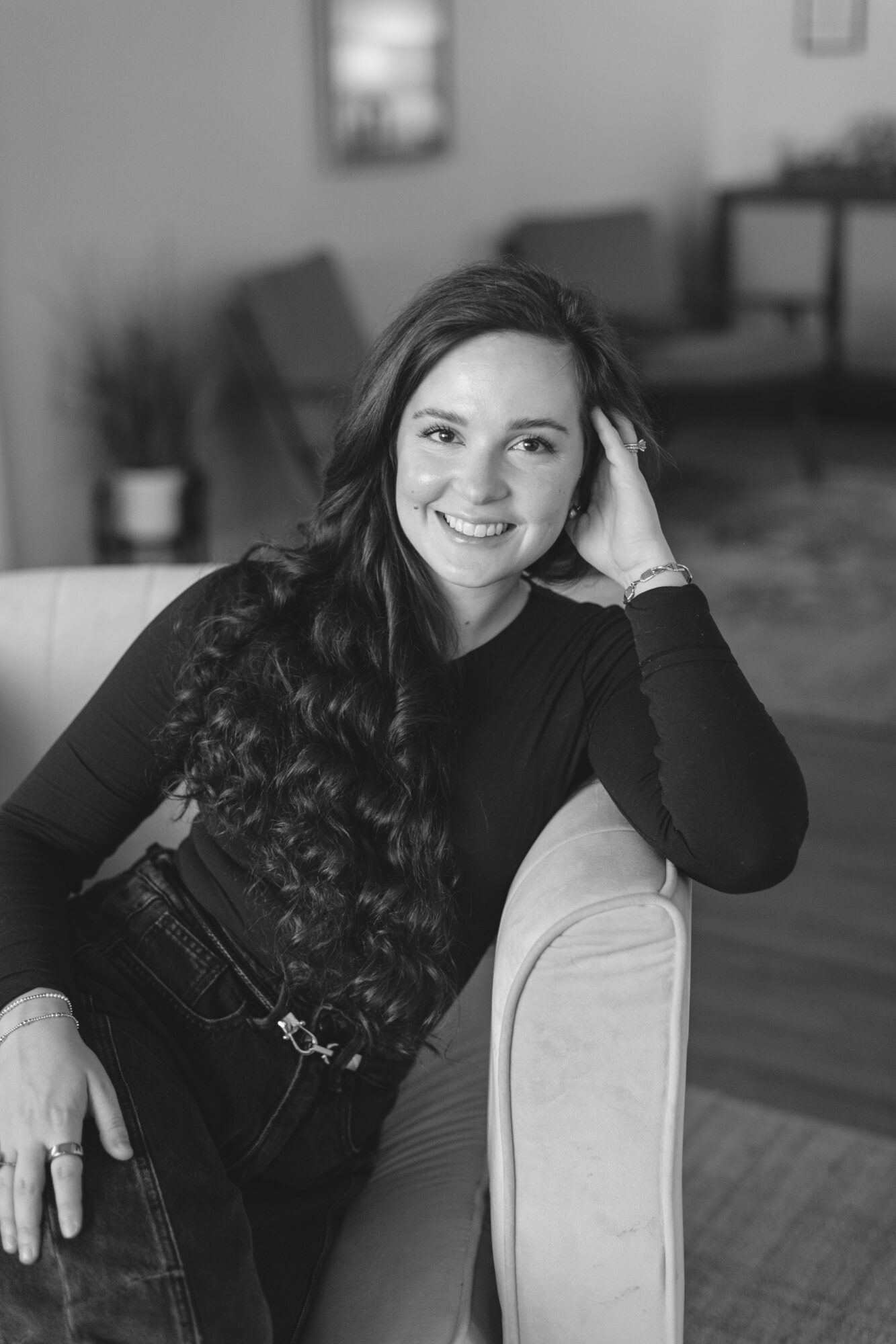 Smiling woman with long wavy hair sitting on a chair in a living room, resting her head on her hand.