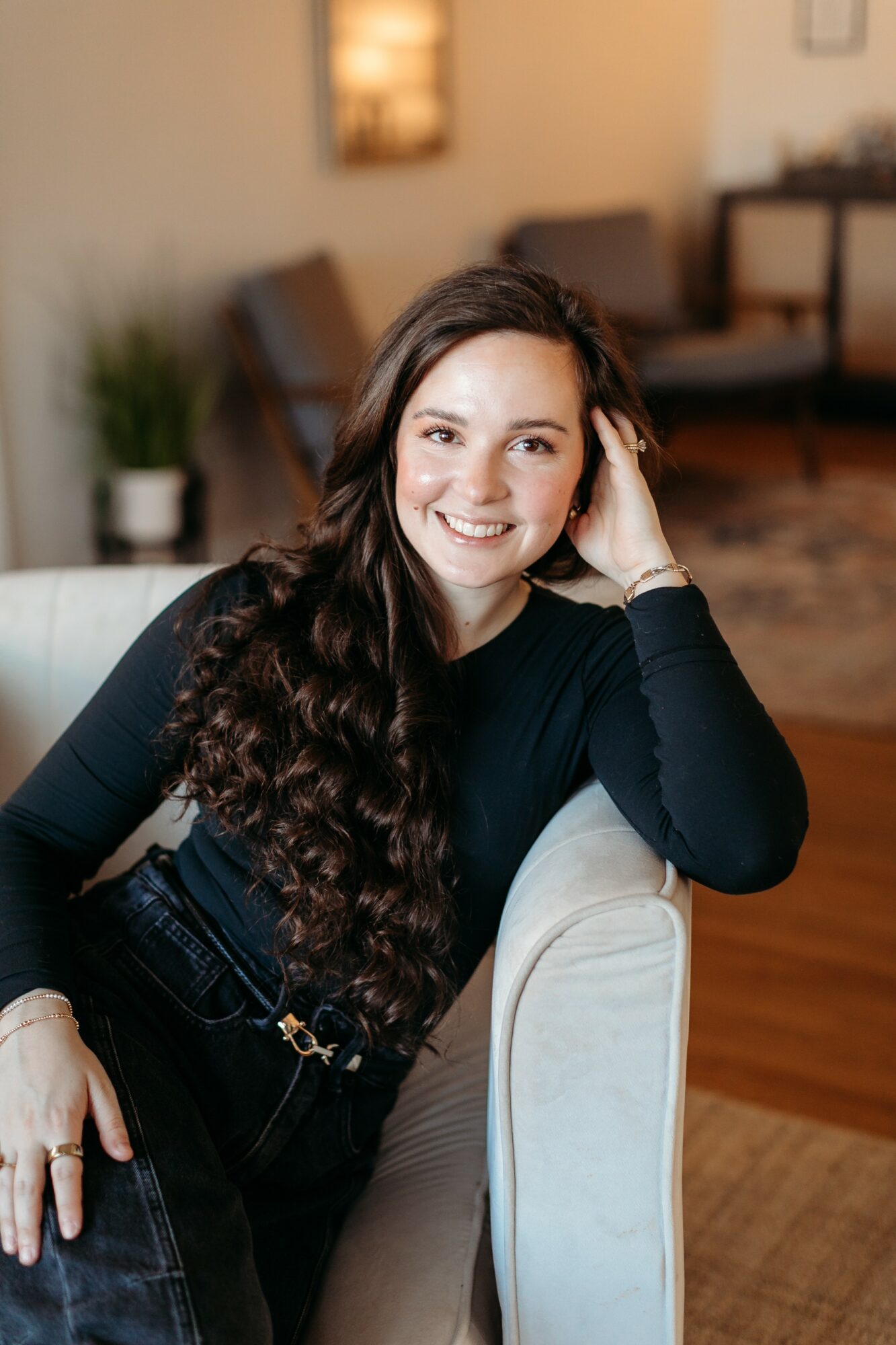 Young woman with long, curly hair smiling, sitting on a light-colored chair in a cozy room.
