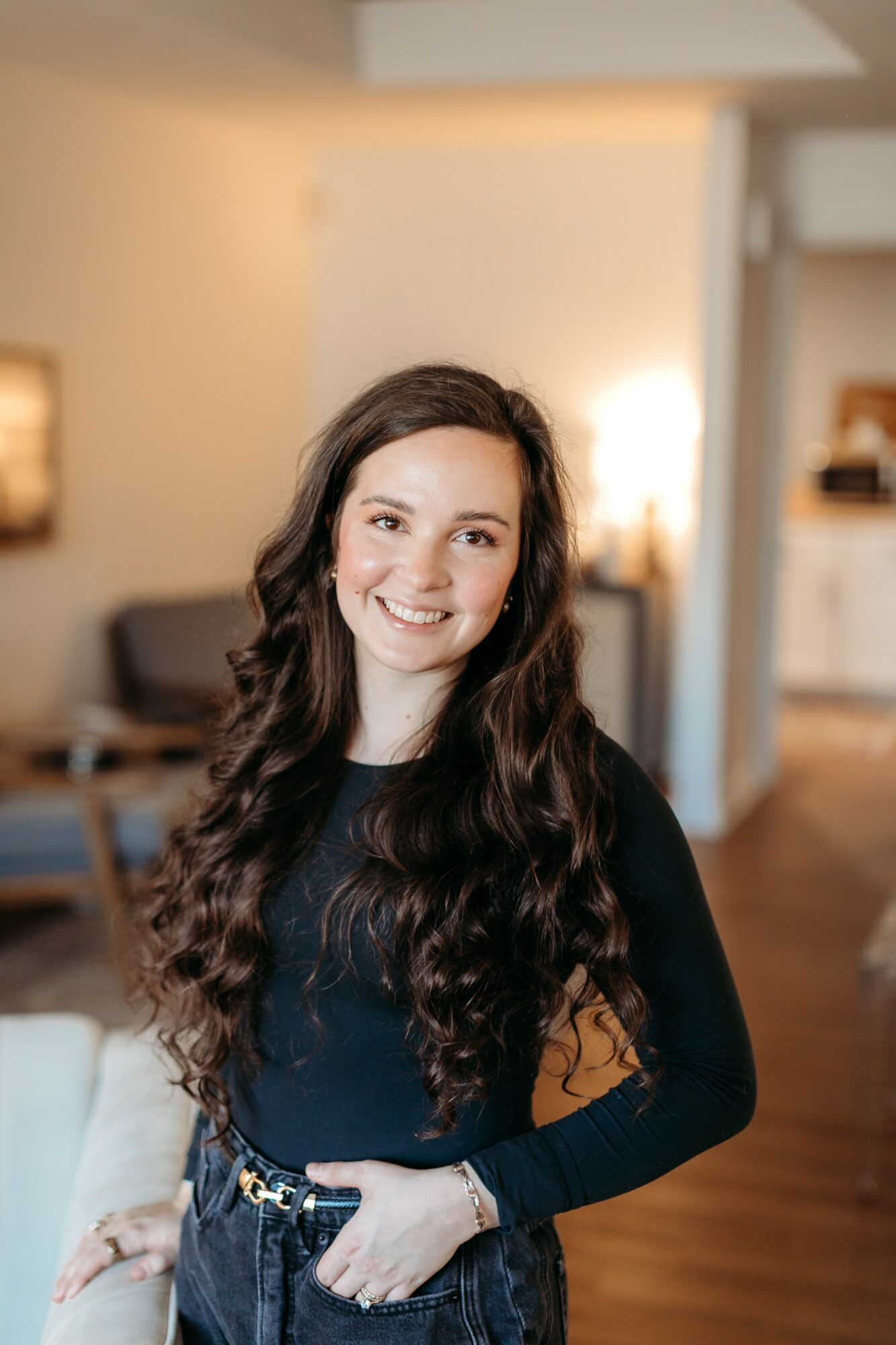 Young woman with long, wavy dark hair smiling indoors, wearing a black top and jeans, in a cozy living room.