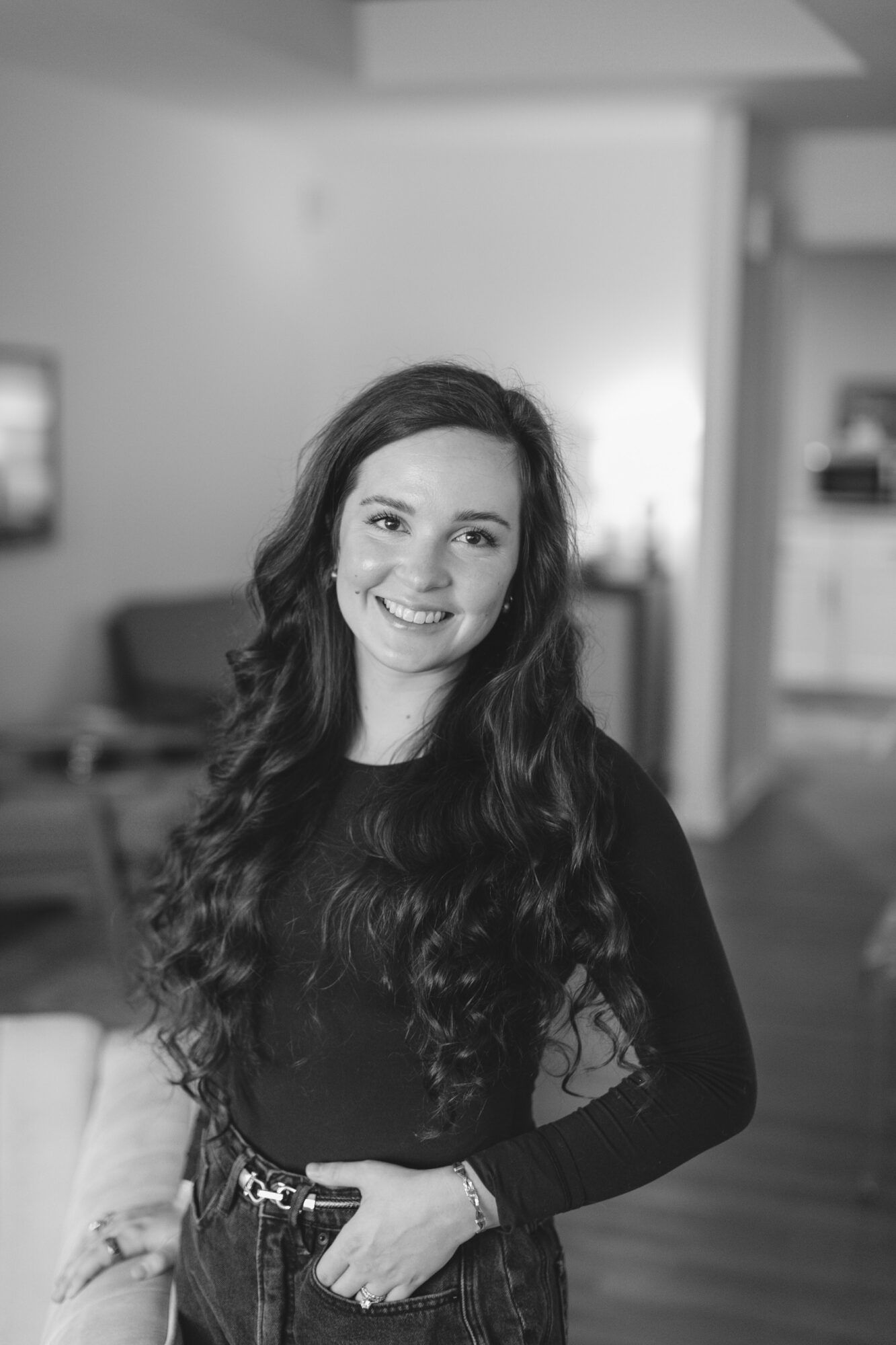 Young woman with long, wavy hair smiling indoors, wearing a dark top and patterned skirt, in a modern room.