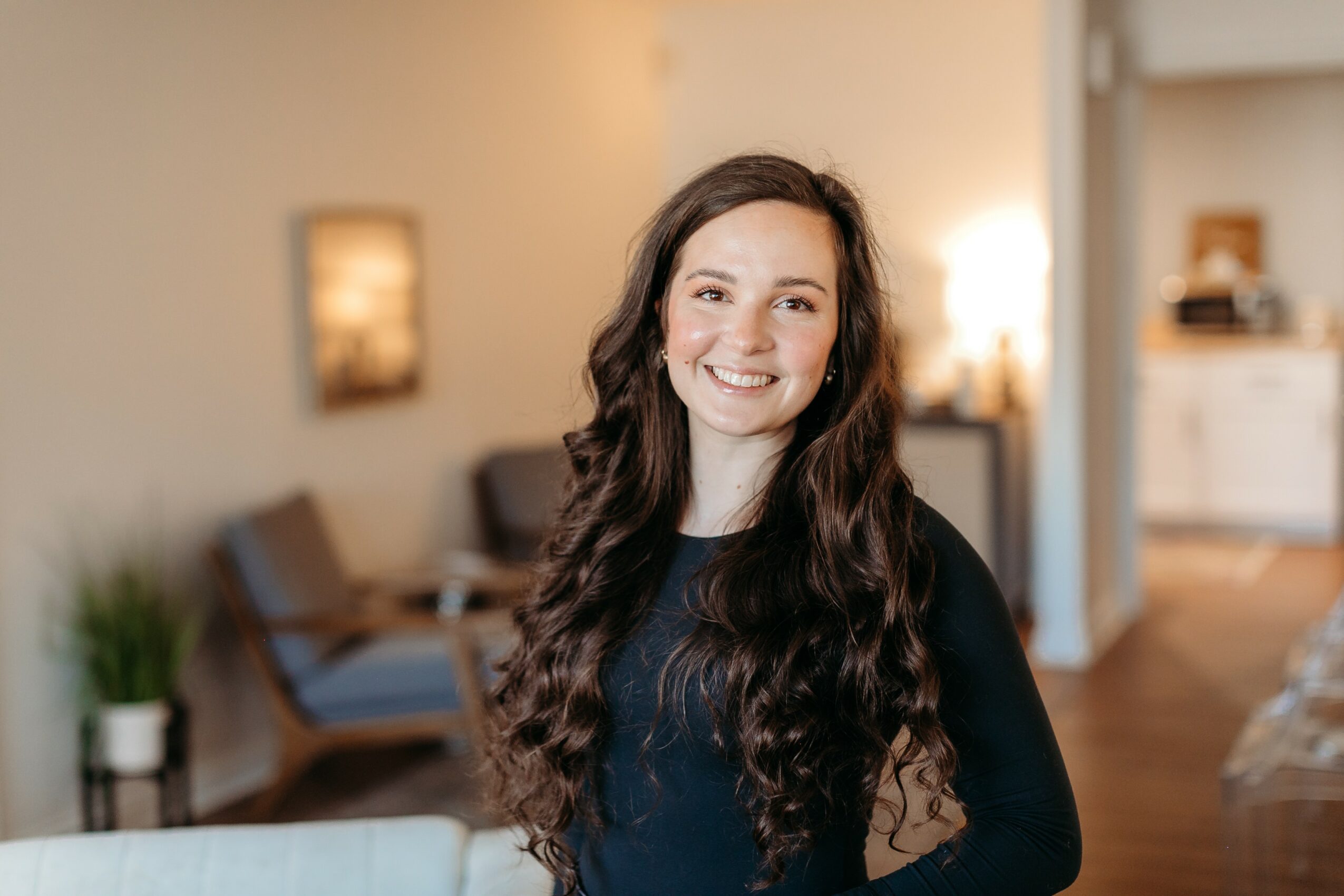 Young woman with long, wavy dark hair smiling in a cozy living room, background includes a sofa, plants, and artwork.