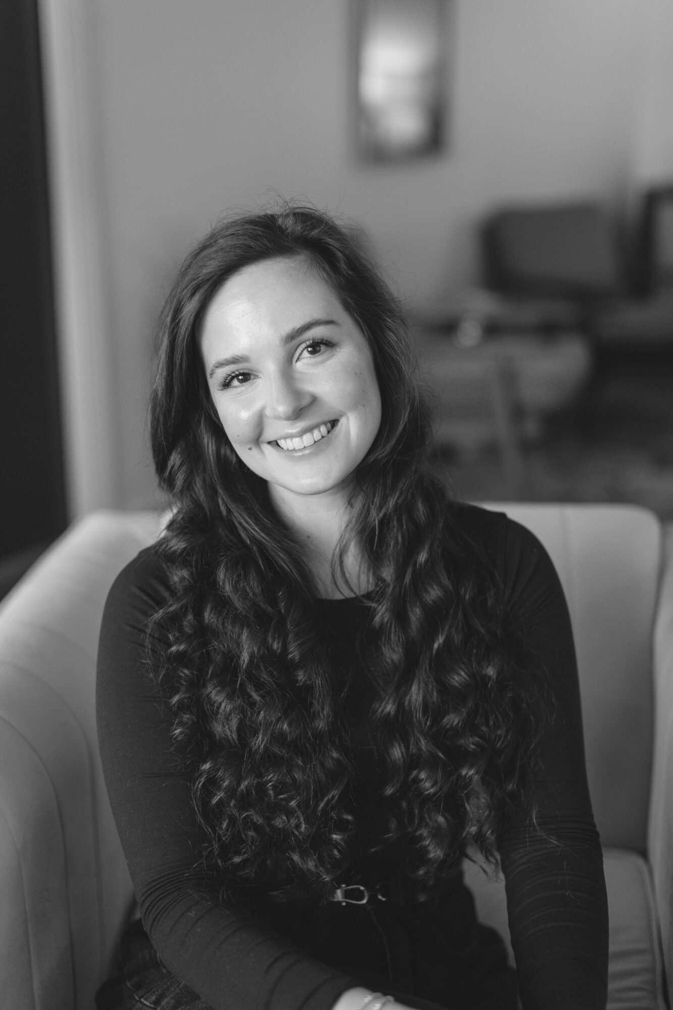 Smiling woman with long wavy hair sitting indoors, background with furniture, in black and white.