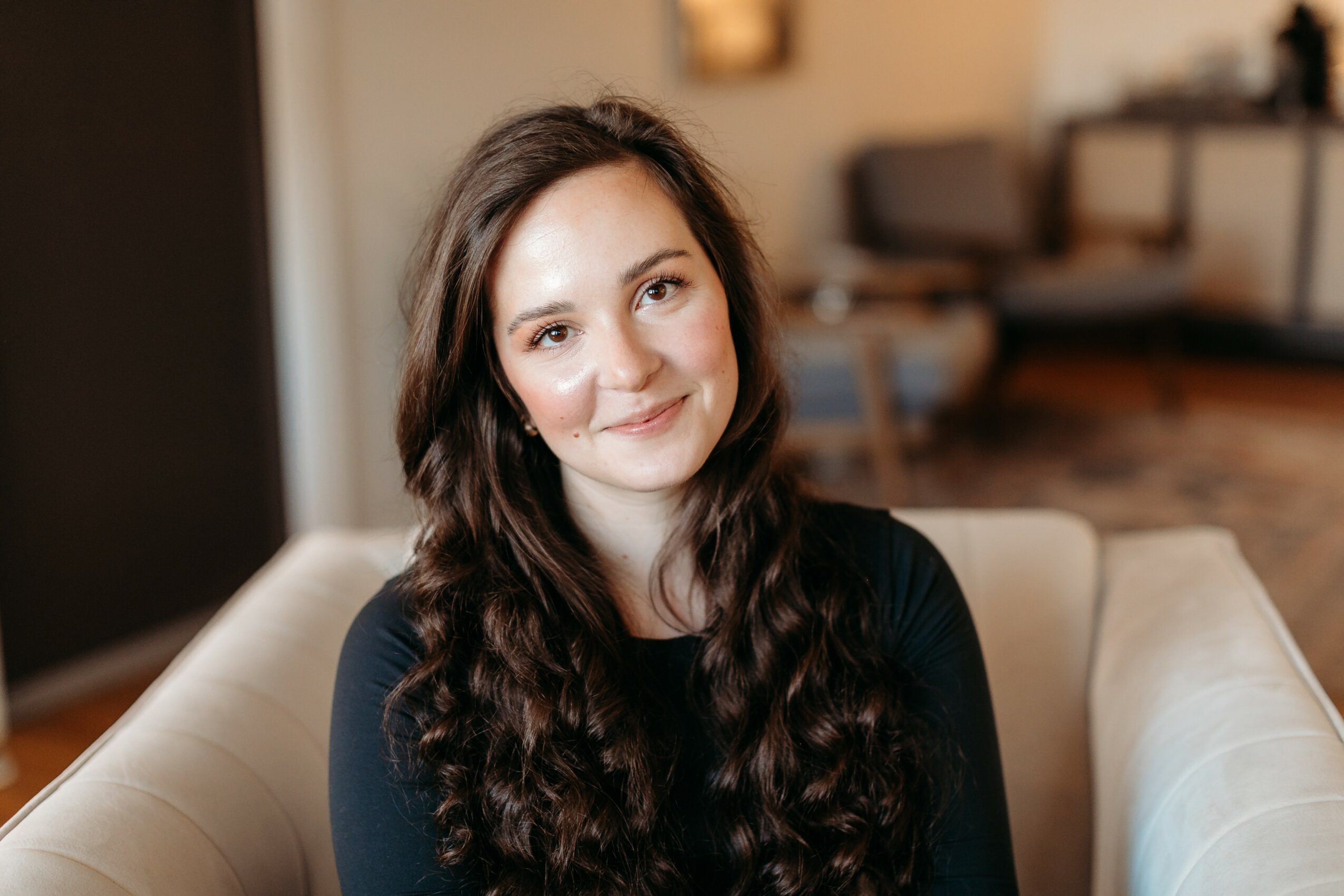 Young woman with long, wavy dark hair smiling while sitting on a beige sofa in a cozy living room.