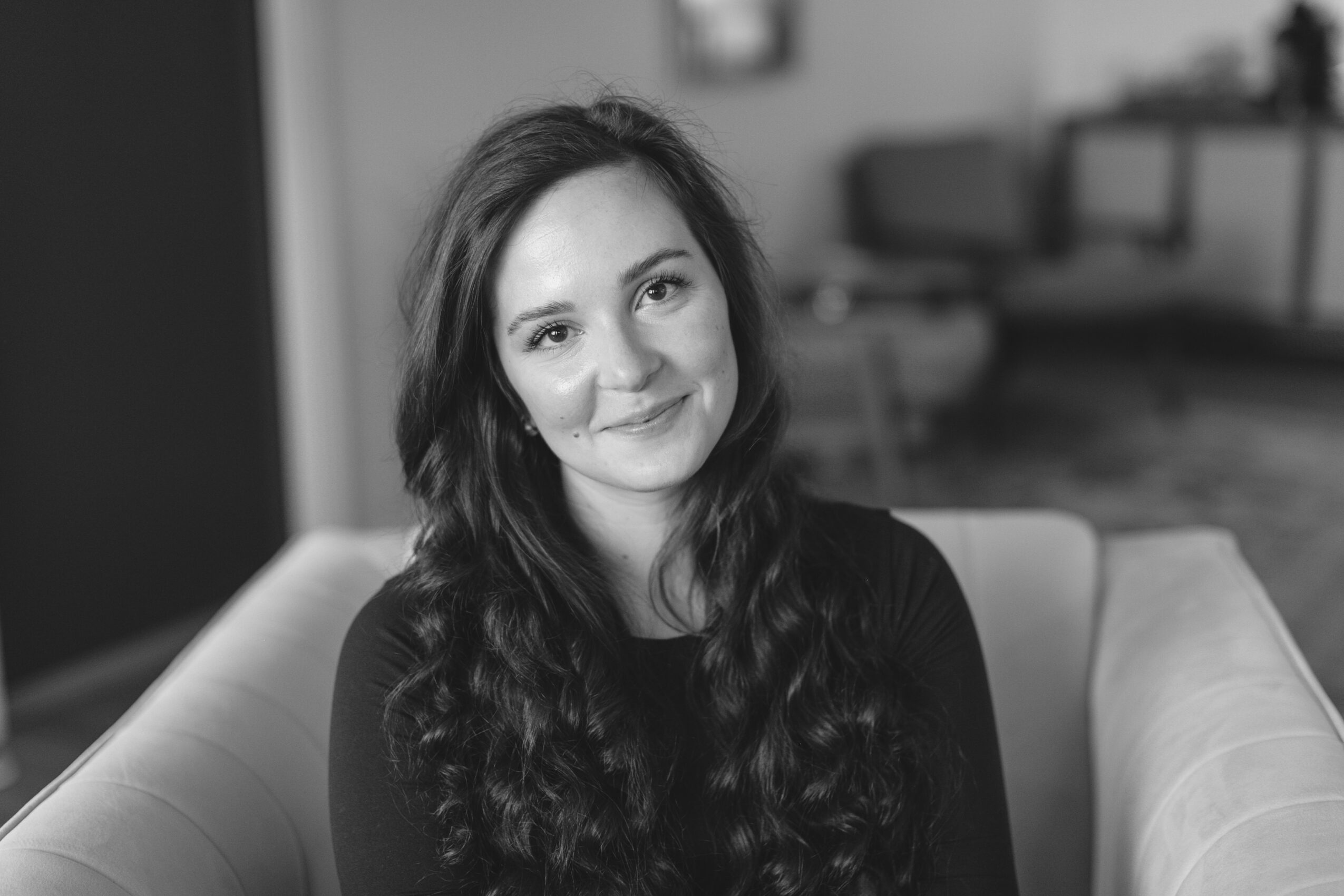 Young woman with long wavy hair sitting on a sofa, smiling, in a living room, black and white photo.