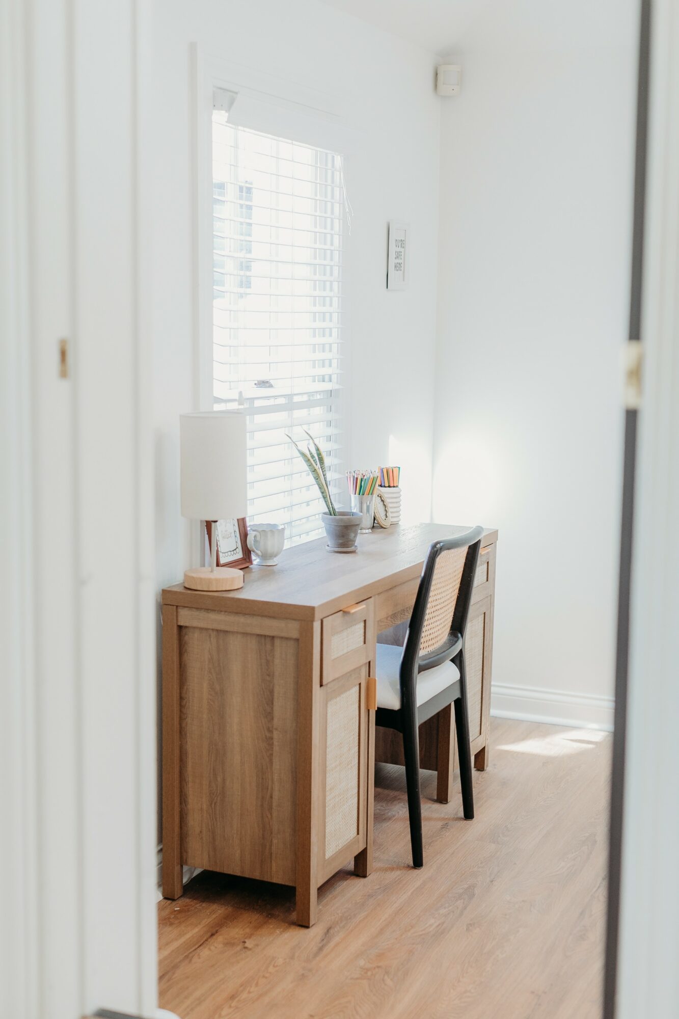 Wooden desk with a chair, lamp, and plant near a window in a bright room.