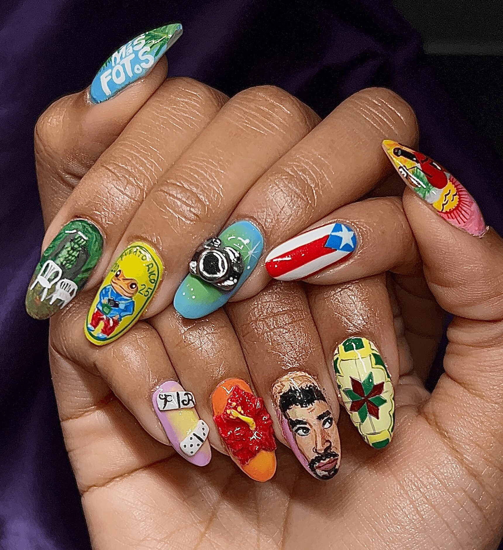 Close-up of hands with colorful, detailed nail art featuring various designs including a camera, superhero symbols, a woman, and abstract patterns.