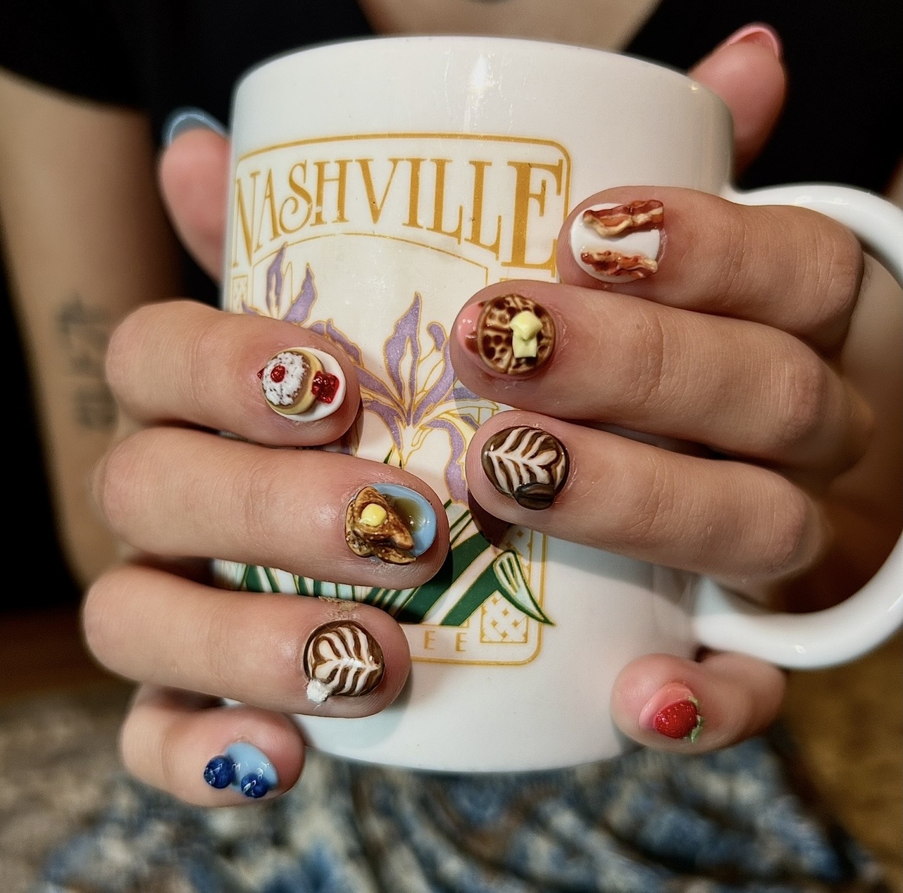 Person holding a Nashville-themed mug with decorated fingernails, some with floral and gold designs, others with stripes and solid colors.