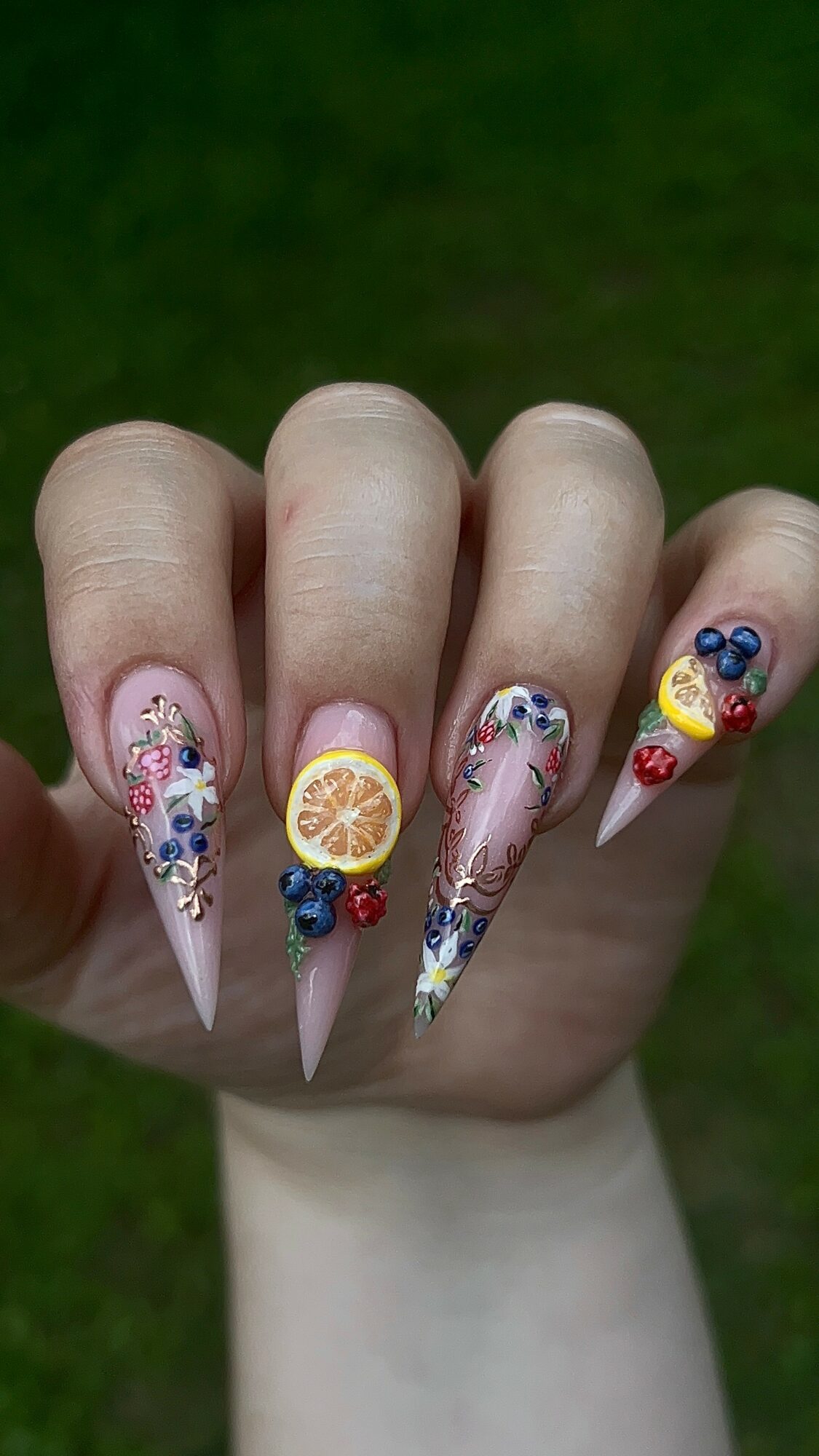 Hand with long, decorated nails featuring fruit and floral designs, set against a green background.