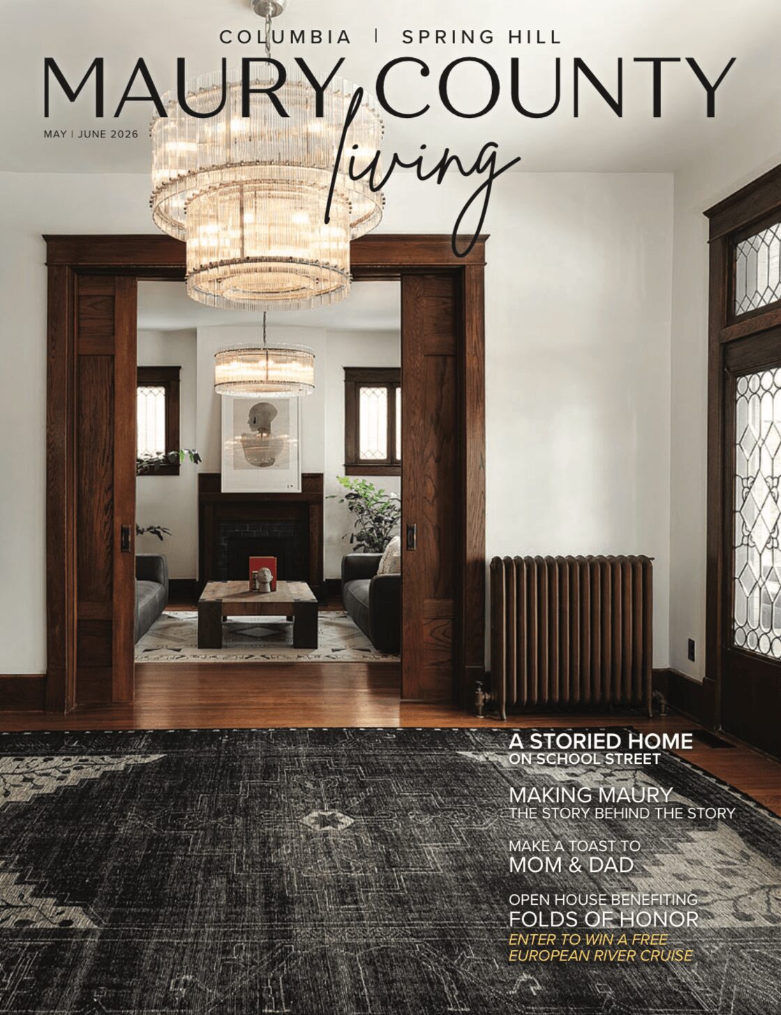 Living room with chandelier, wooden doorway, fireplace, and large windows, featuring a dark rug and hardwood floors.