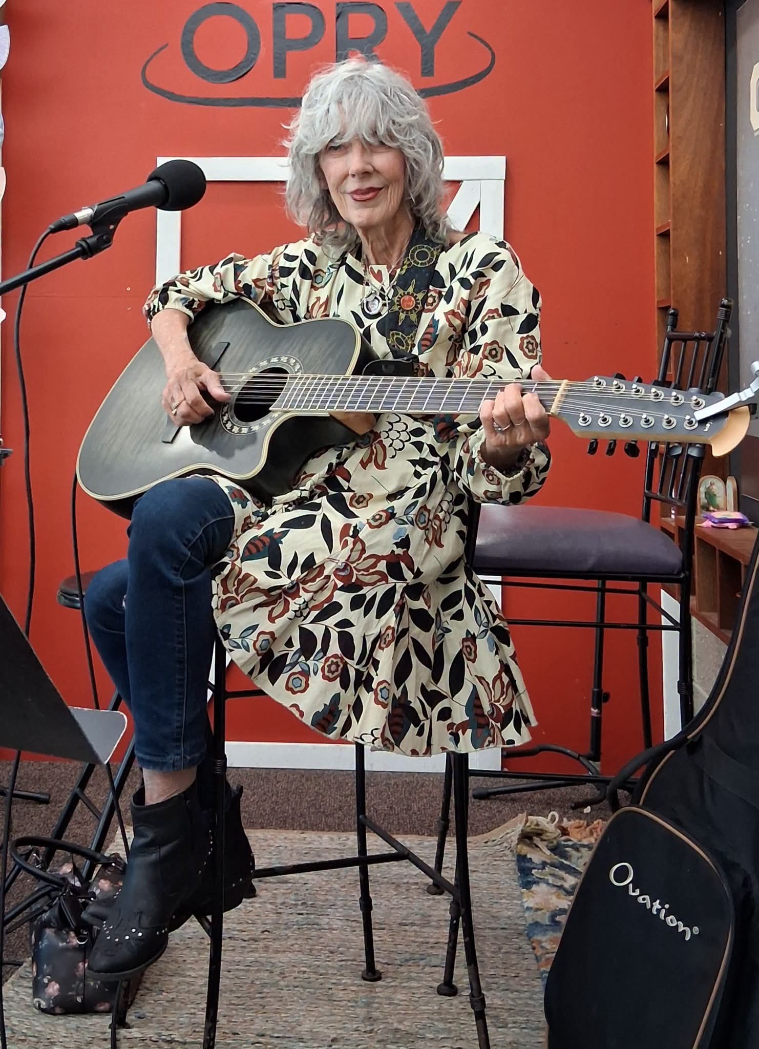Woman with gray hair playing an acoustic guitar on stage, seated on a chair, with a red background and microphone nearby.