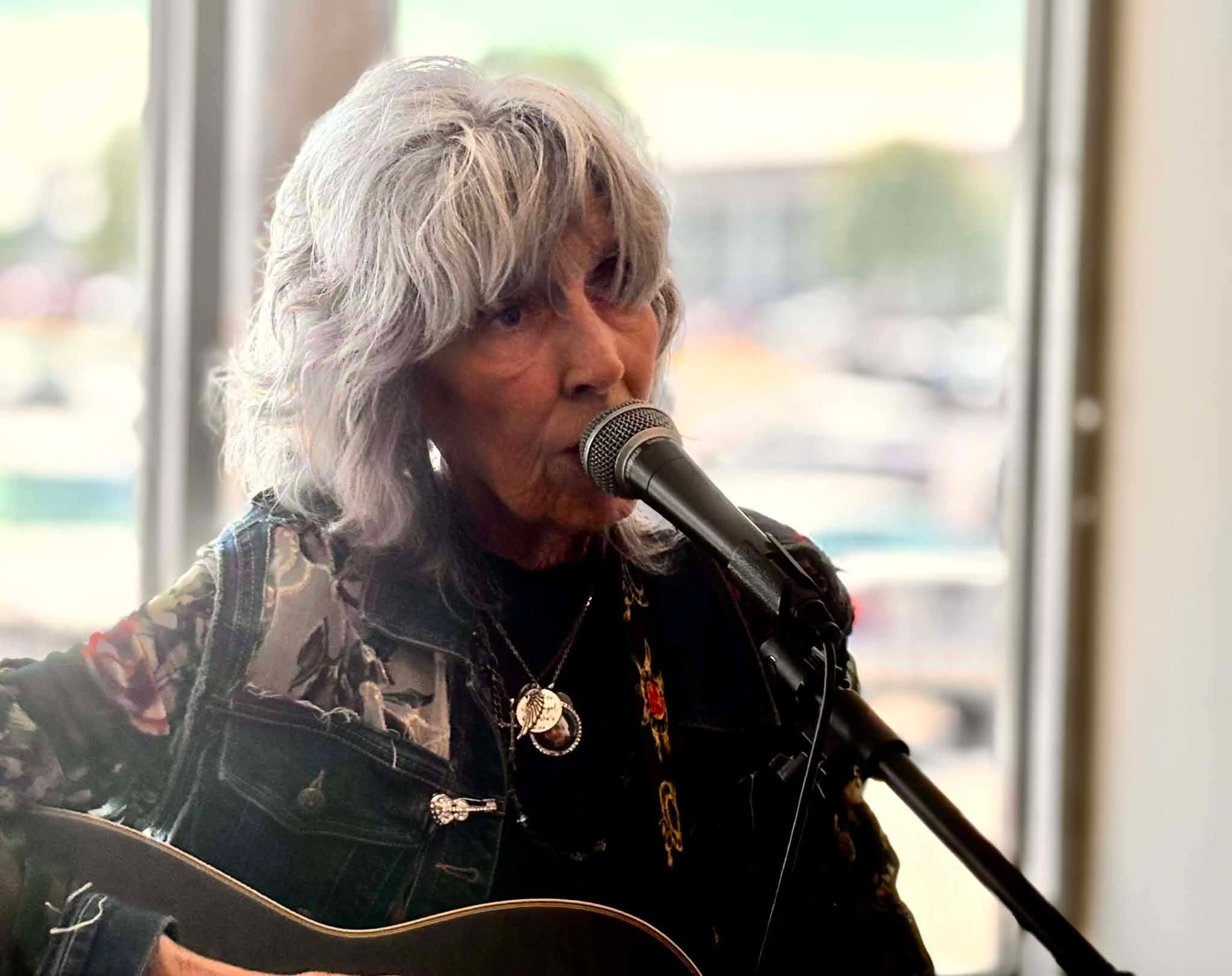 Older woman with gray hair singing into a microphone while playing guitar, indoors with large windows behind her.