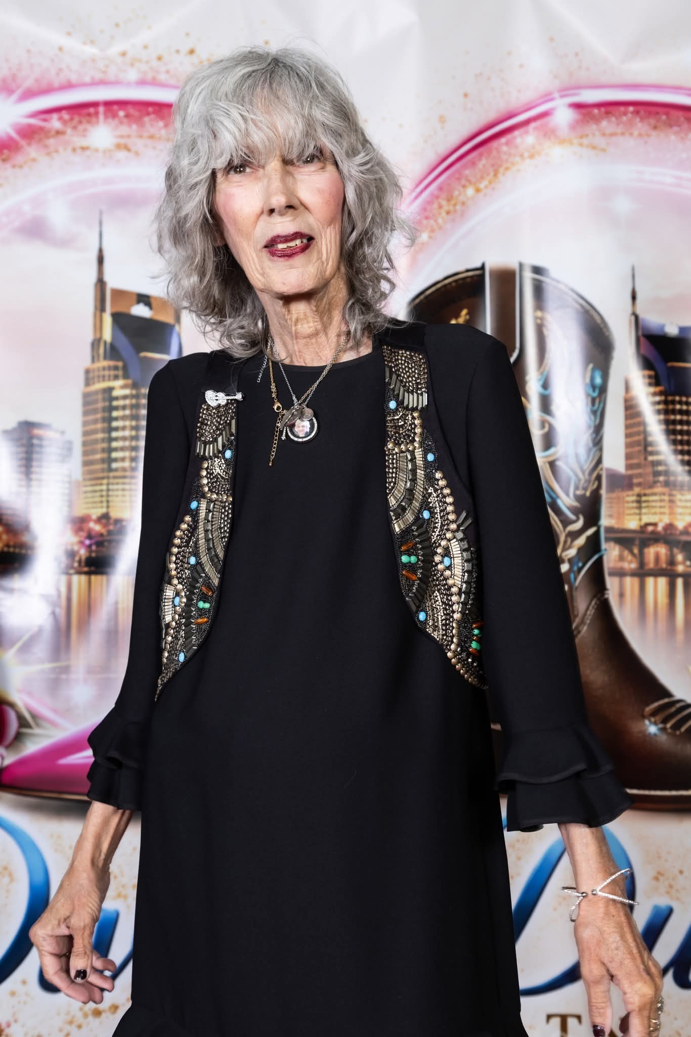 Older woman with gray hair wearing a black dress and a decorated jacket, standing in front of a cityscape backdrop.