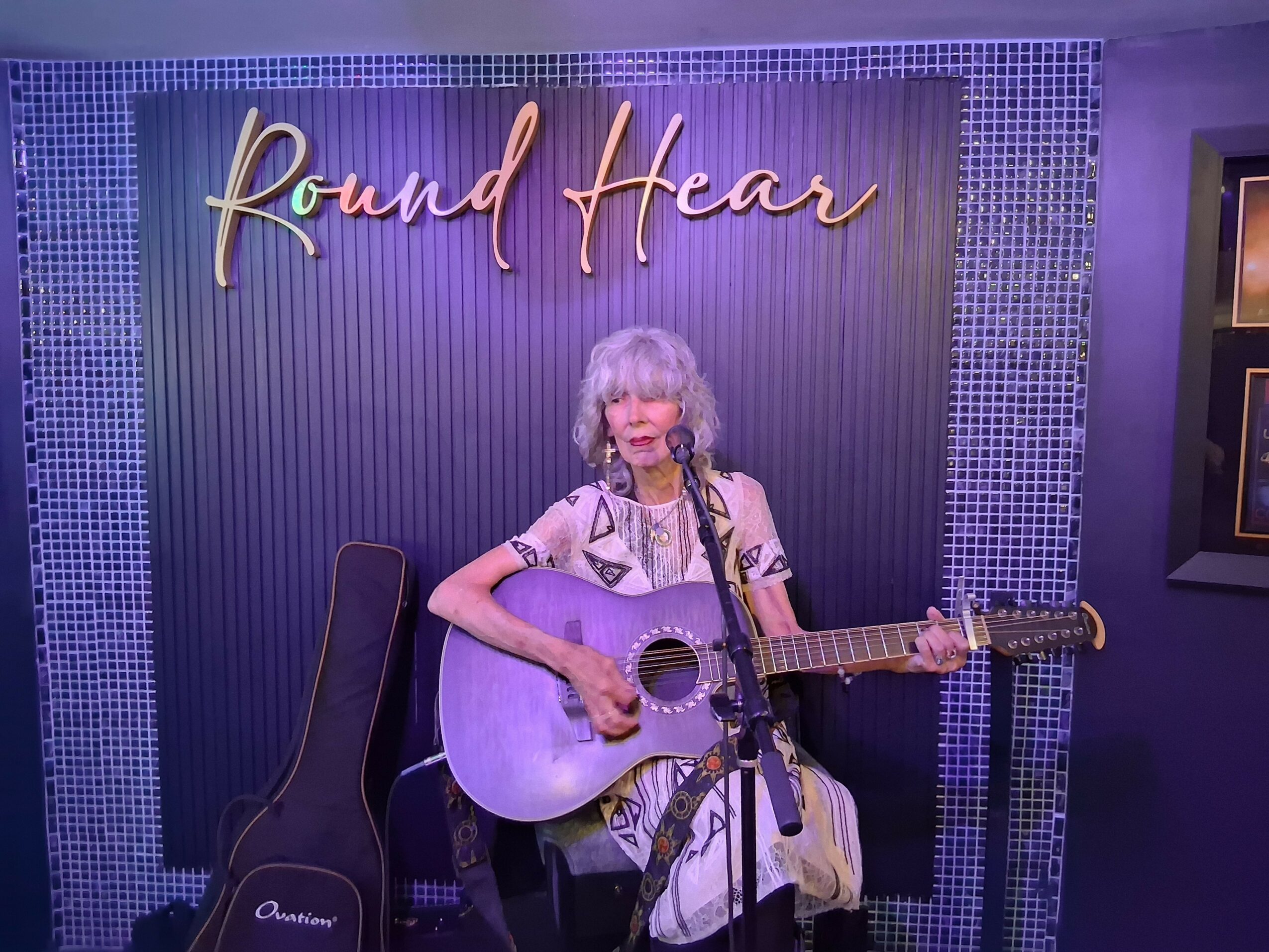 A woman with curly gray hair plays guitar and sings into a microphone on stage with a sign reading 'Round Heart' behind her.
