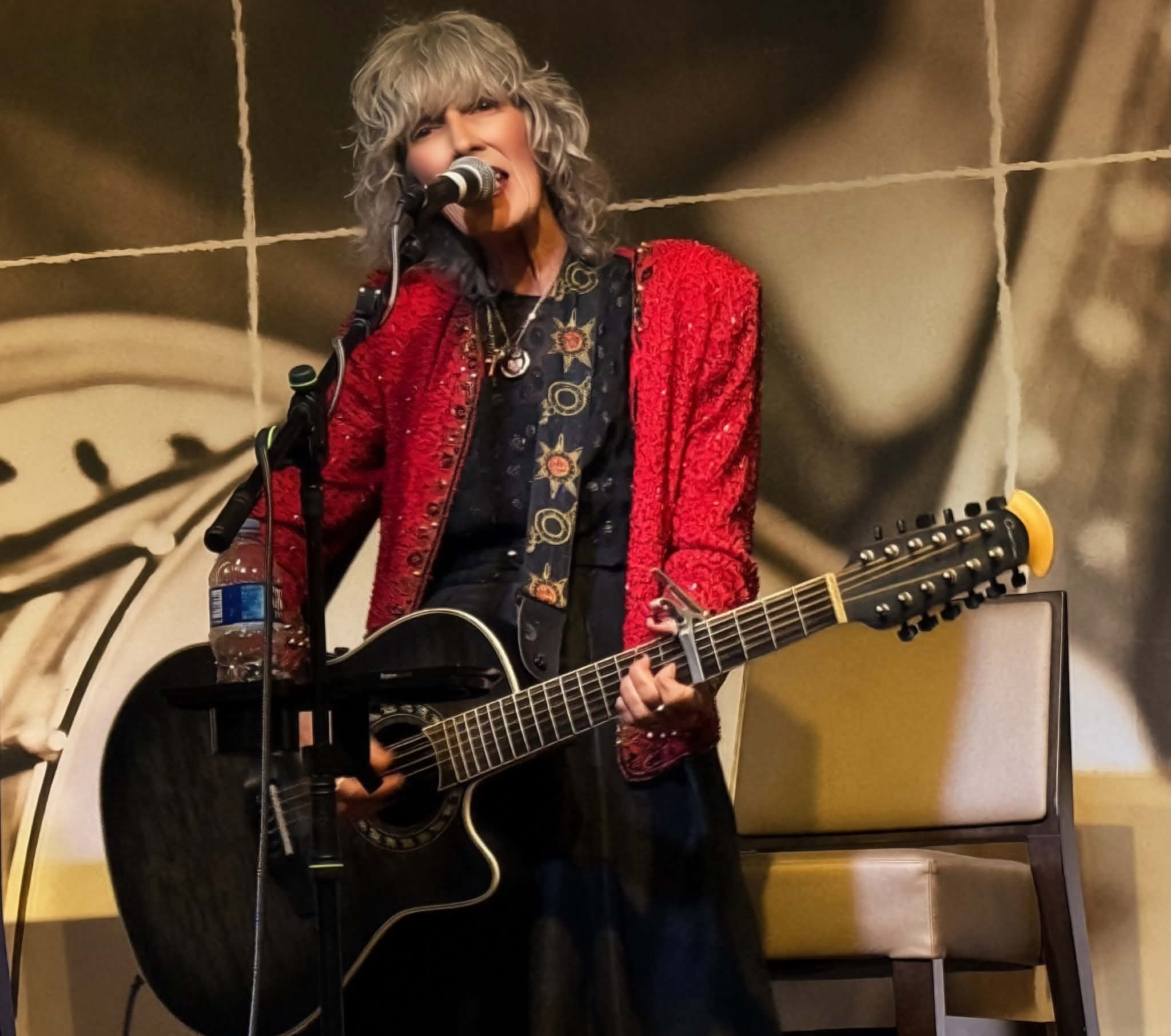 Person with curly gray hair singing into a microphone while playing an acoustic guitar, wearing a red jacket.