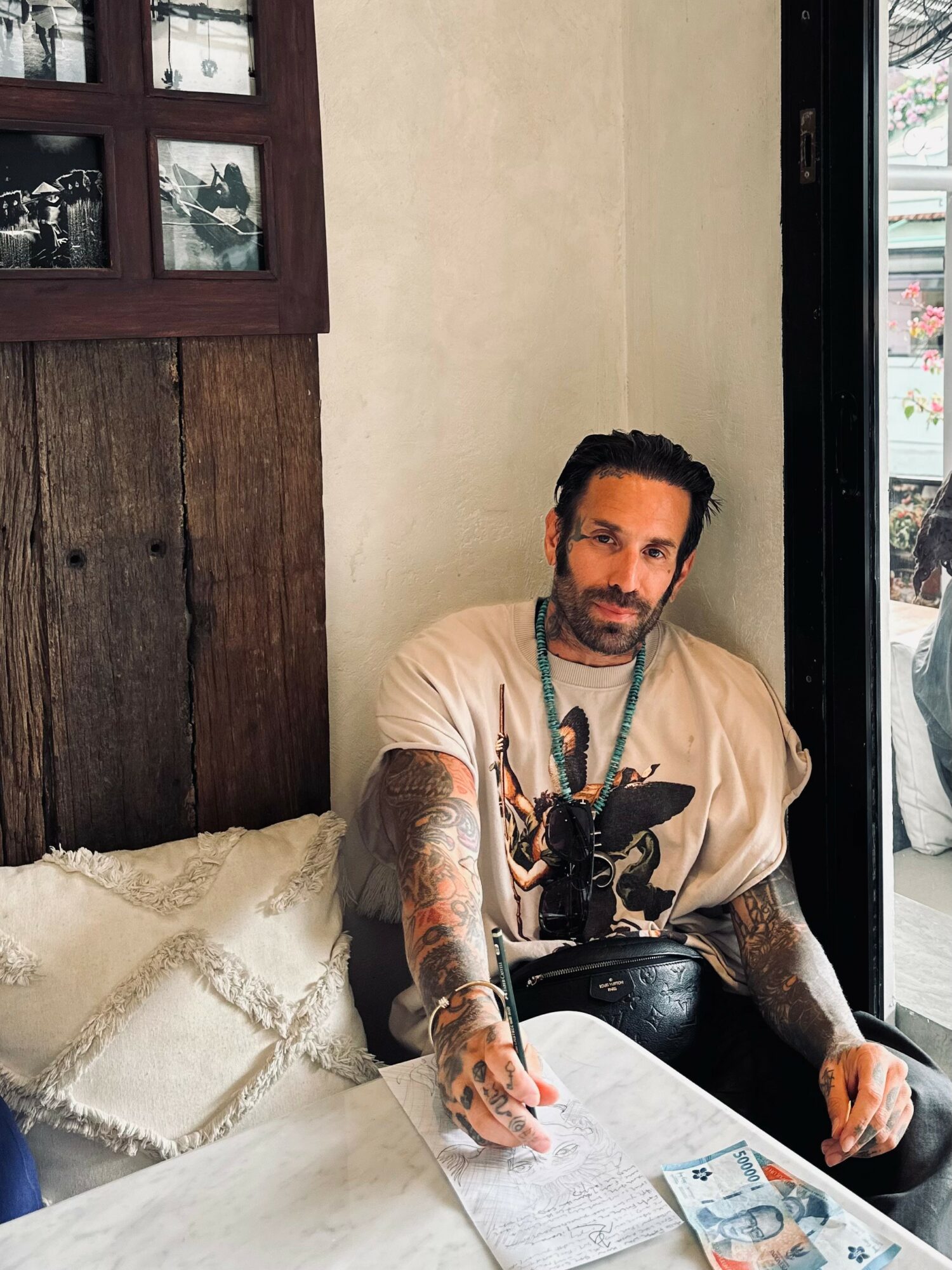 Man with tattoos sitting on a bed, looking at the camera, in a room with wooden and beige walls, near a window.