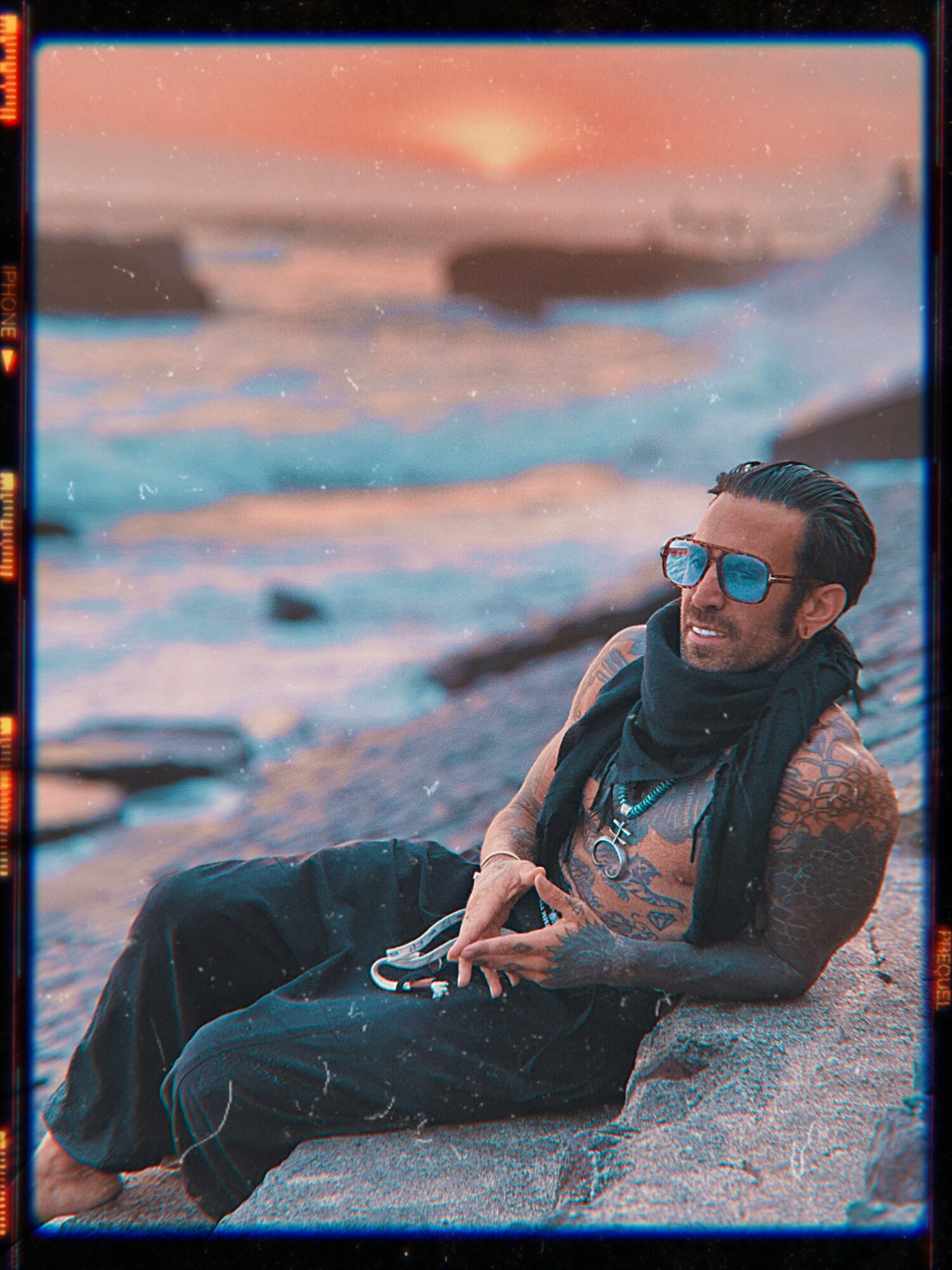 Man with sunglasses and tattoos relaxing on rocks near the ocean at sunset.
