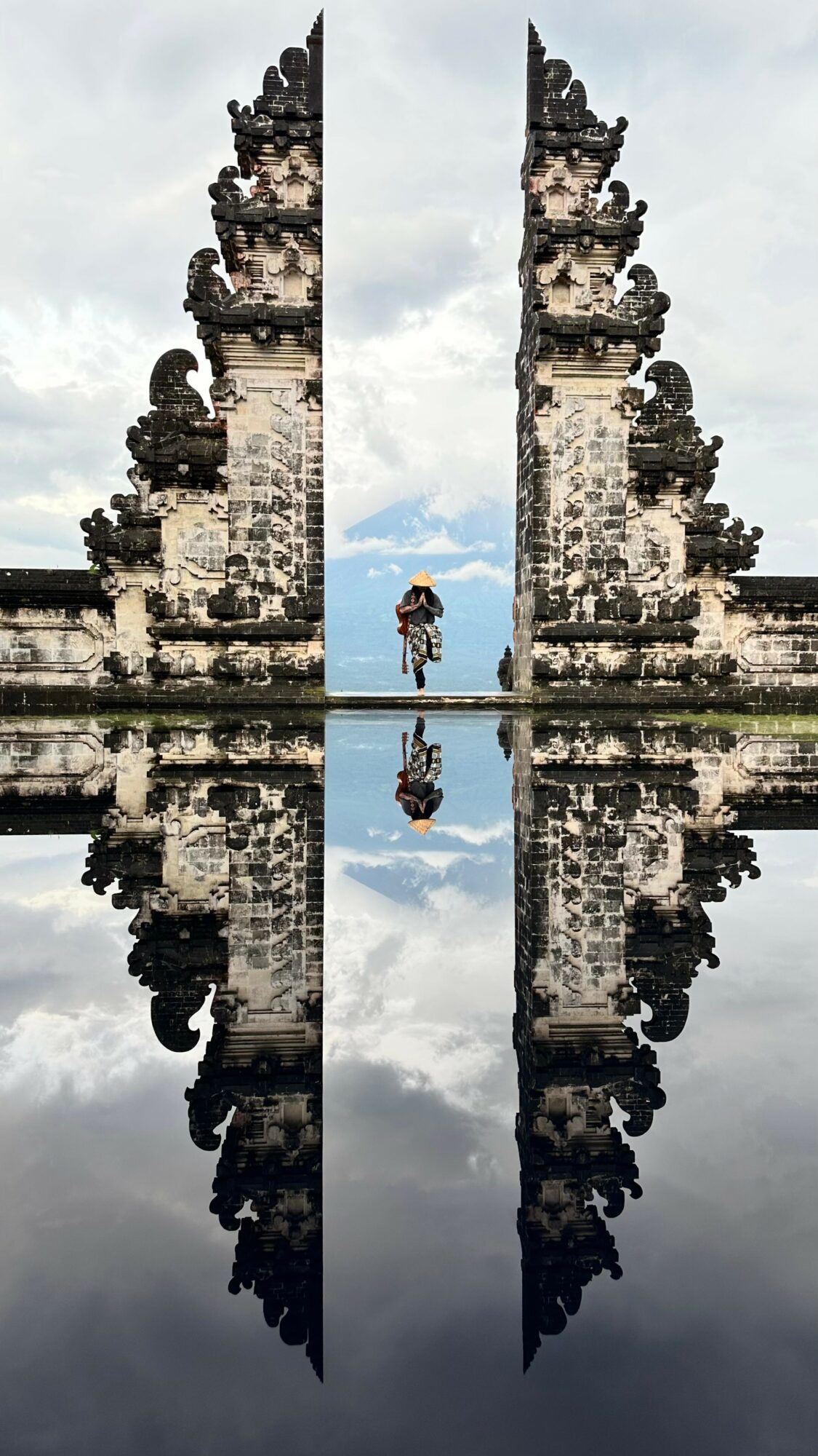 Stone gate with intricate carvings reflected in water, person walking through, cloudy sky background.