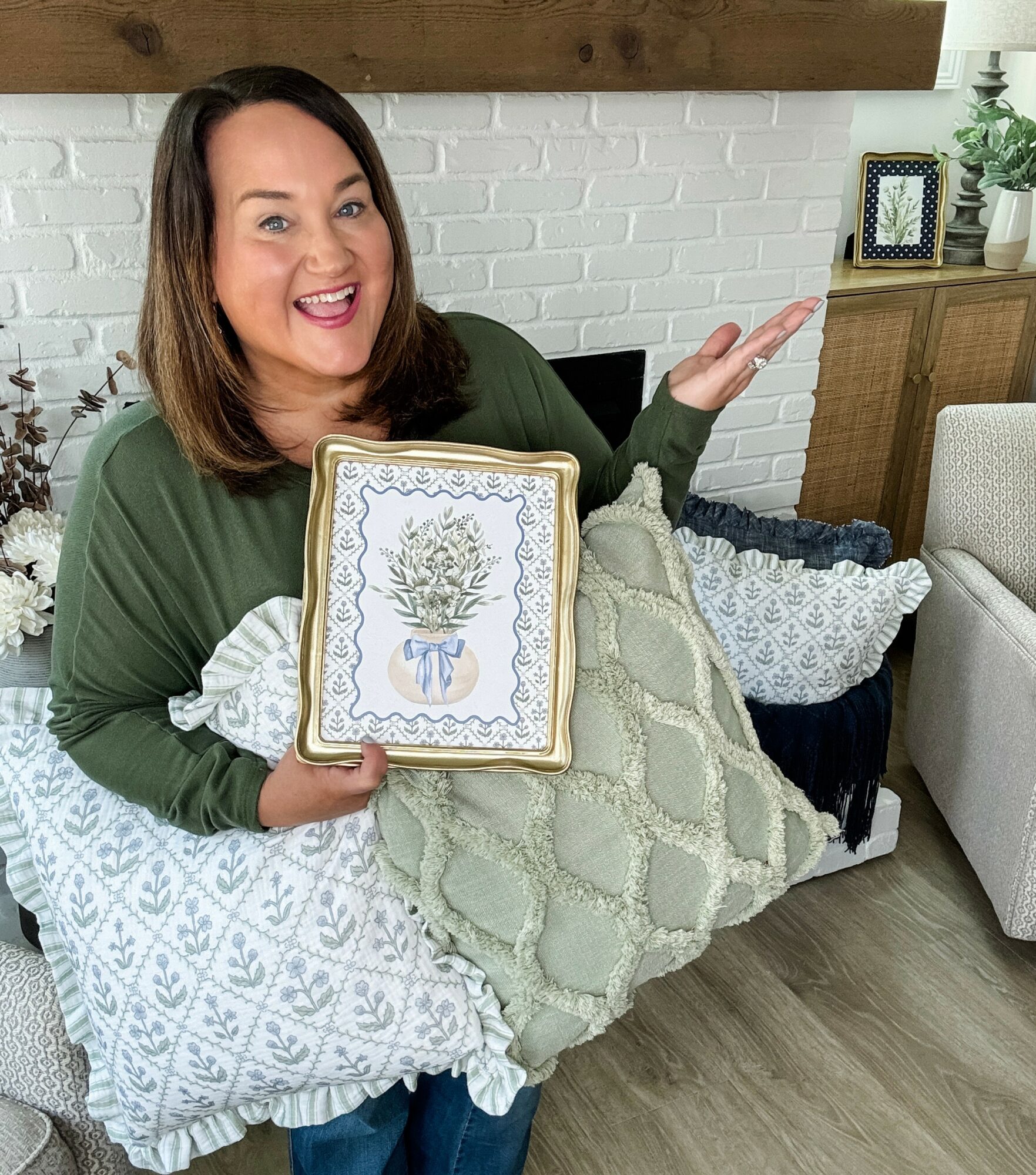Woman smiling, holding a framed picture of a plant, surrounded by pillows in a cozy living room.