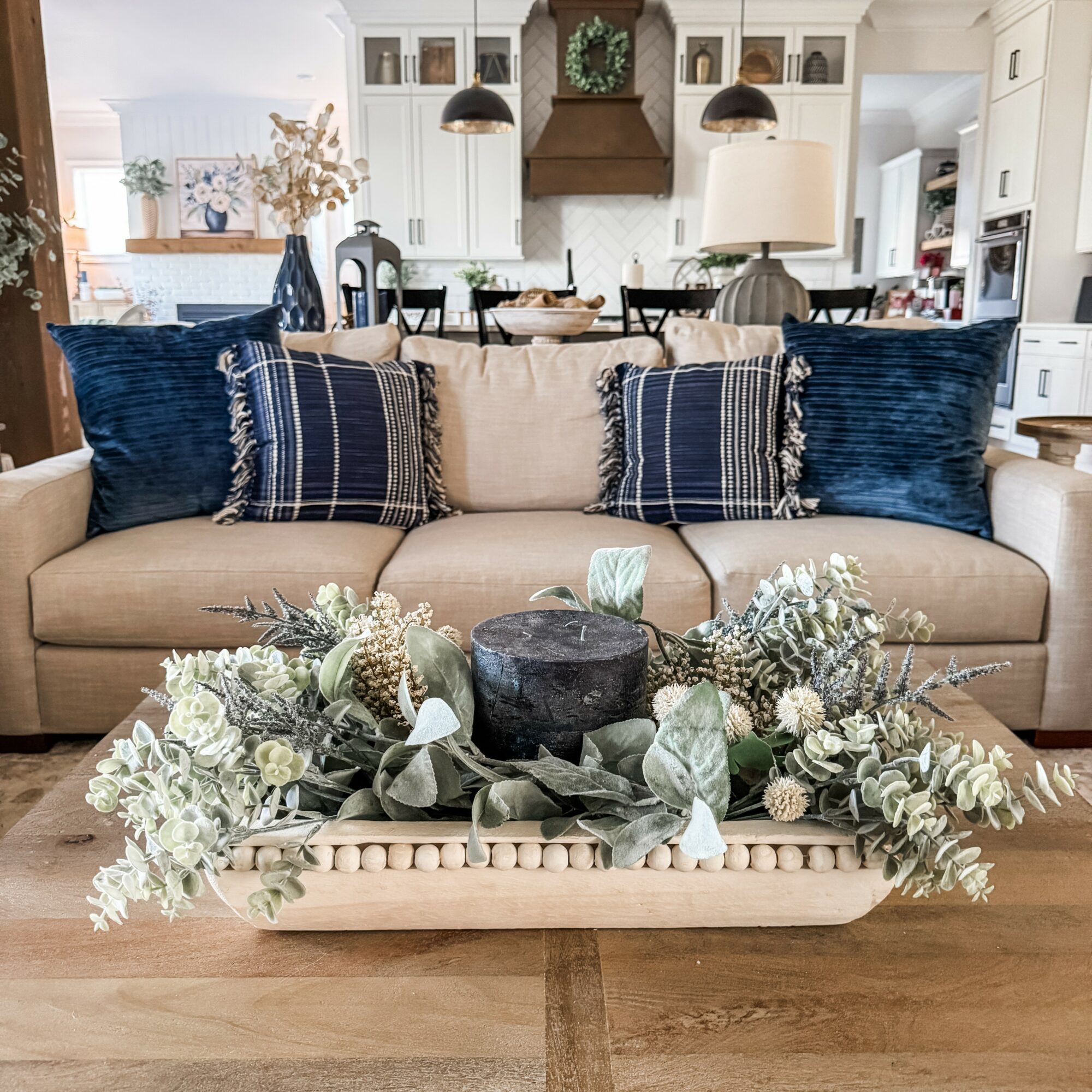 Living room with beige sofa, decorative pillows, and a floral arrangement on a wooden table, kitchen in background.