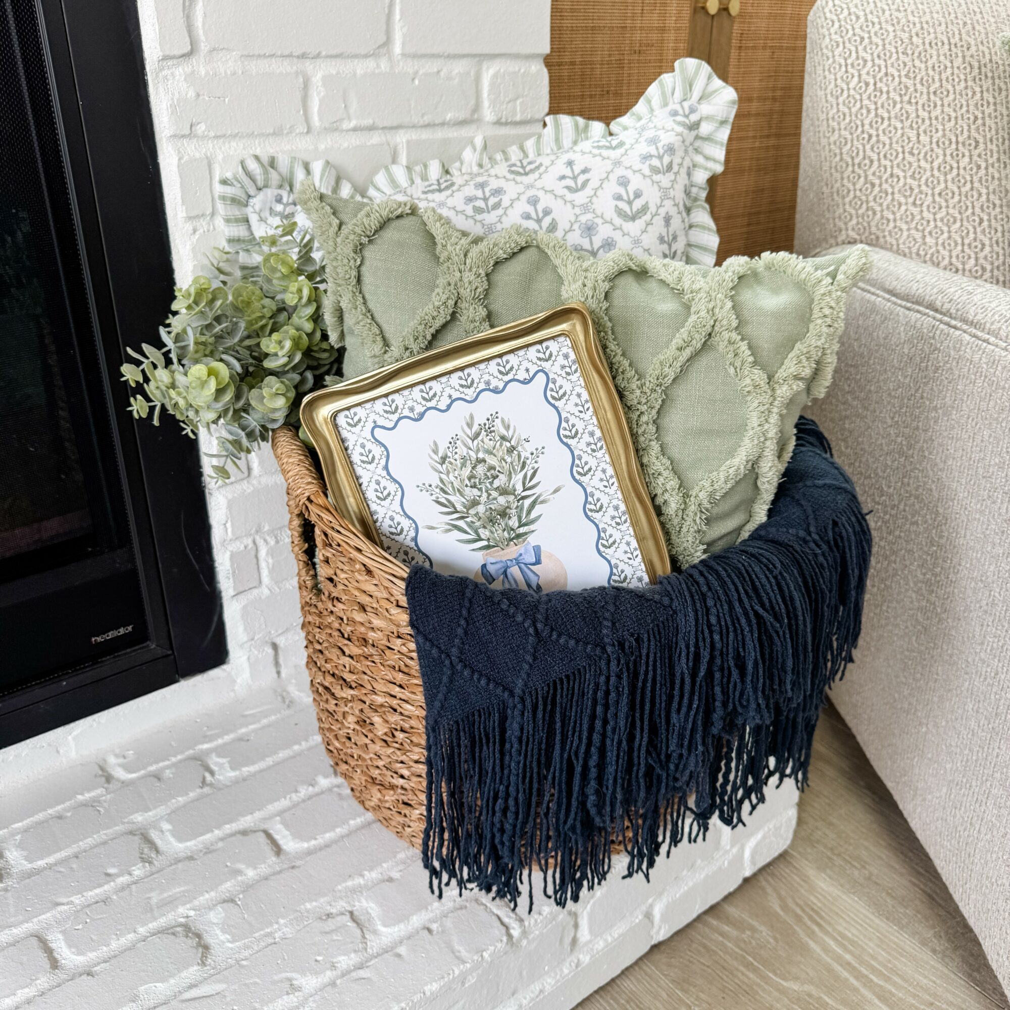 Basket filled with pillows, a framed picture, and a plant, placed next to a white brick wall and a beige sofa.
