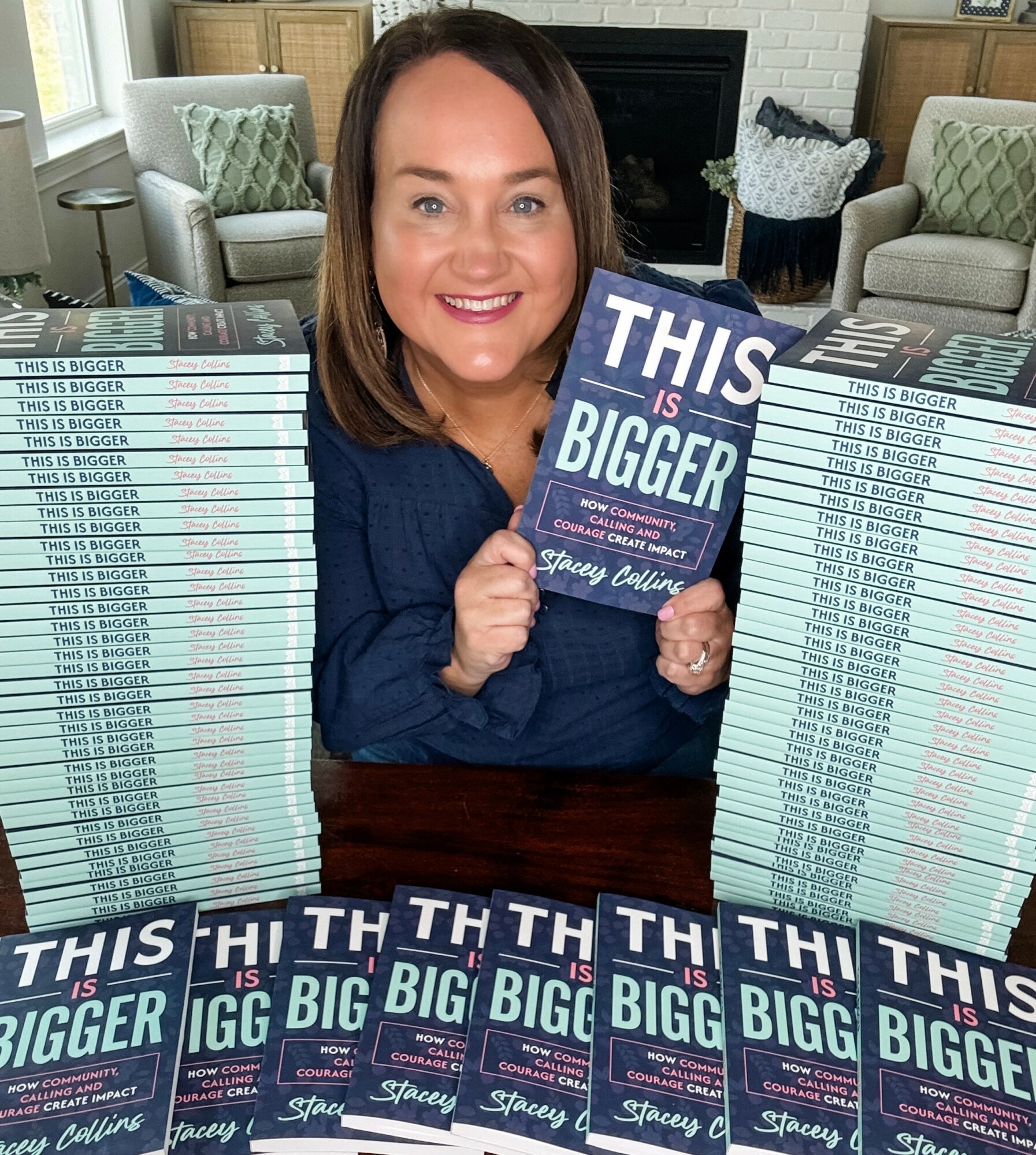 Woman holding a book titled 'This is Bigger' surrounded by stacks of the same book in a living room.