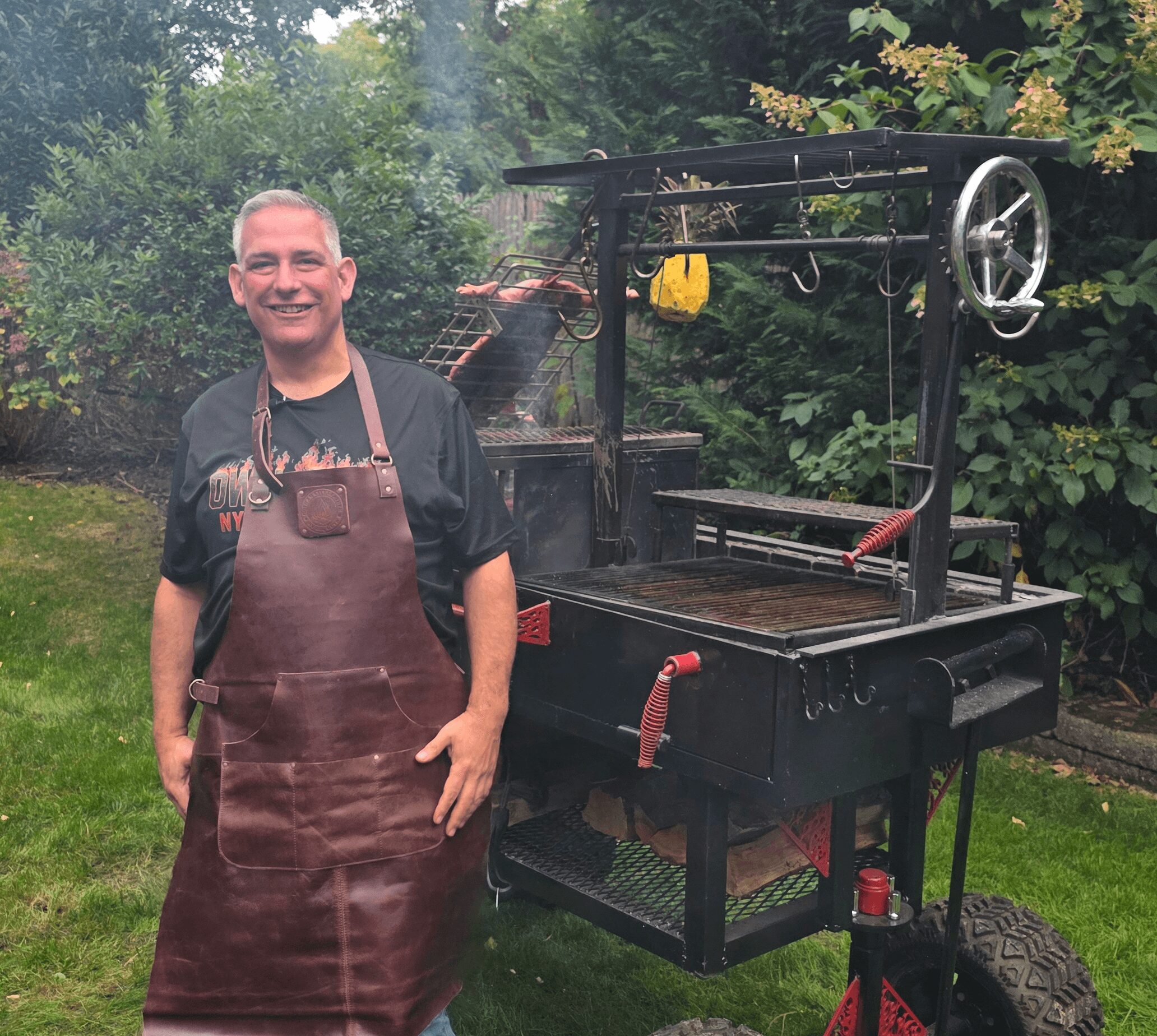 Man wearing apron standing next to a large black barbecue grill outdoors, surrounded by green trees and grass.