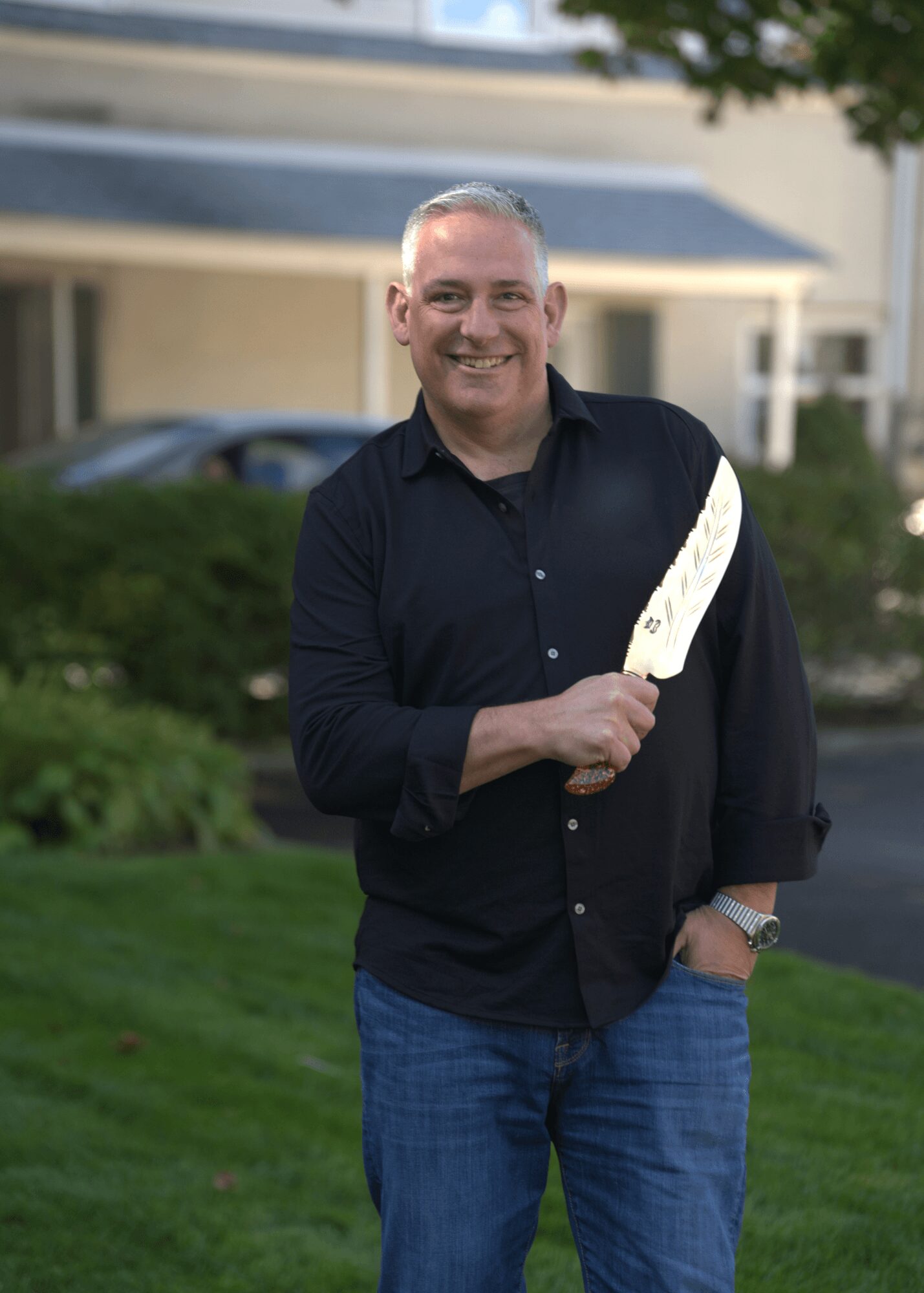 Smiling man in black shirt and jeans holding a white object outdoors, with a house and greenery in background.