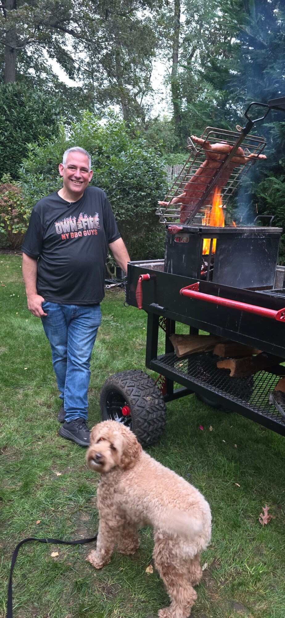Person standing outdoors with a dog on a leash, trees and a grill in the background.