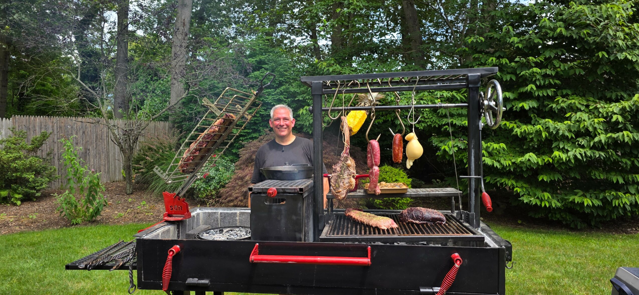 Man standing behind a large outdoor grill with hanging utensils and food, in a backyard with trees and a fence.